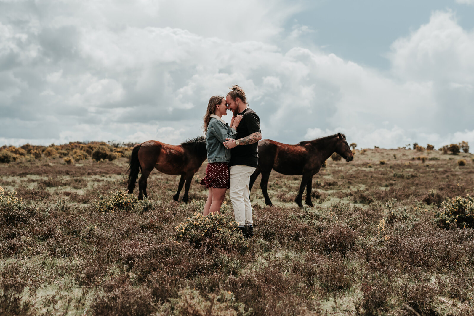 Couple gently embrace in the middle of the stunning moorlands of New Forest, in front of two wild horses