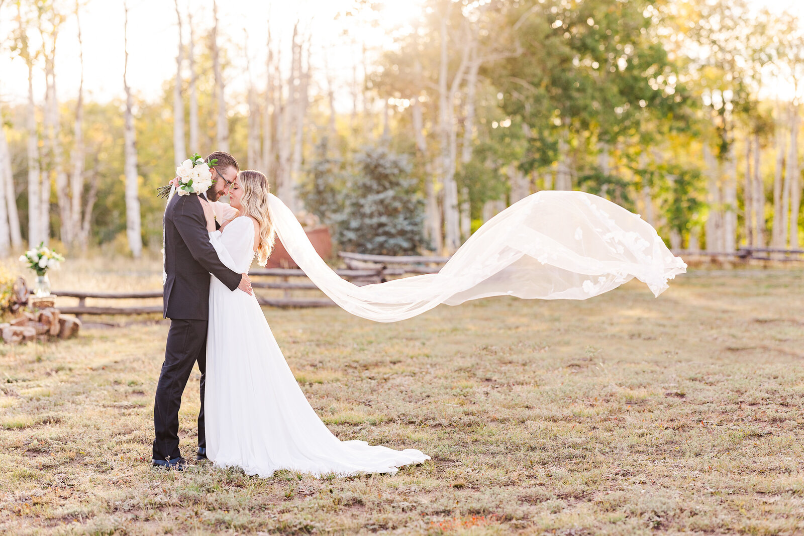 Groom holding bride in field as her veil flies against the sunset