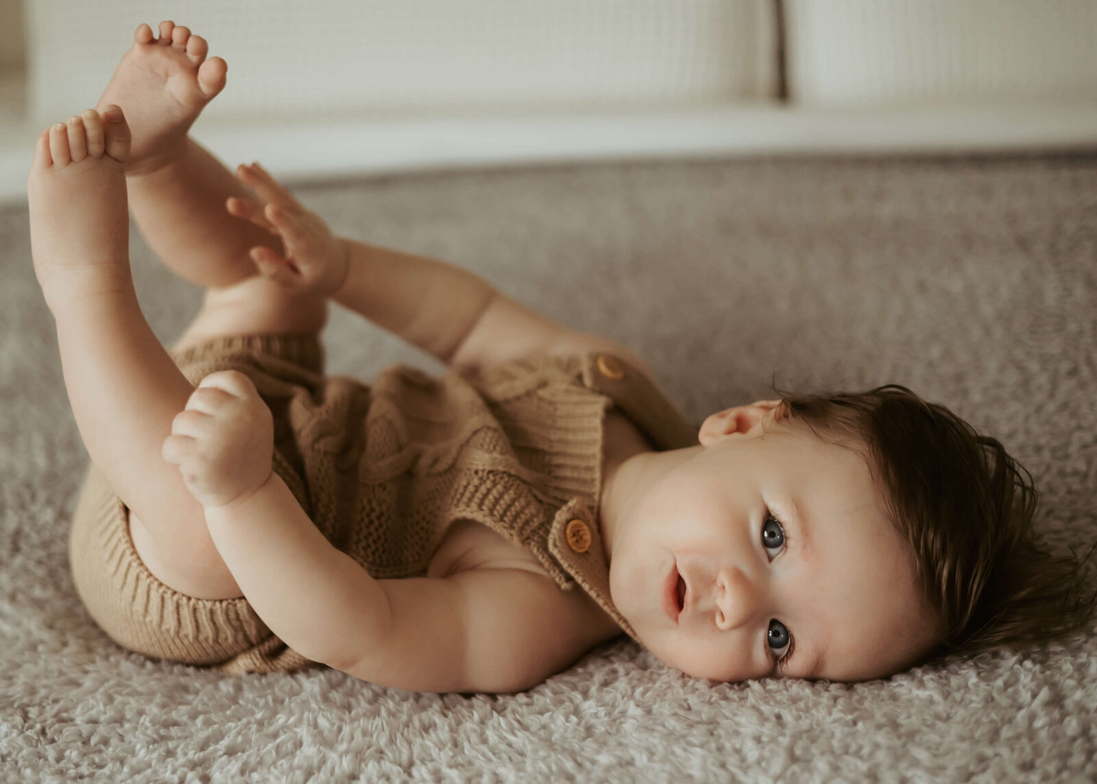A cute baby with blue eyes and brown hair is staring at the camera while on his back for his in-home photography experience.