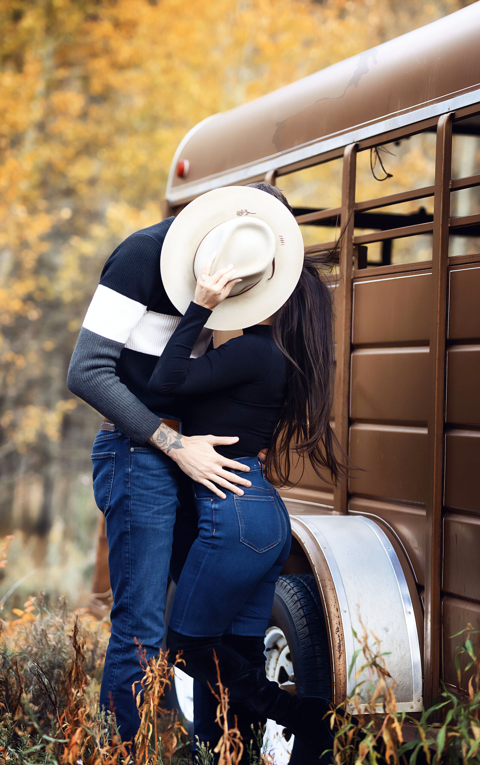An adorable couple kiss behind a cowboy hat, being playful and fun at their Aspen engagement photoshoot.
