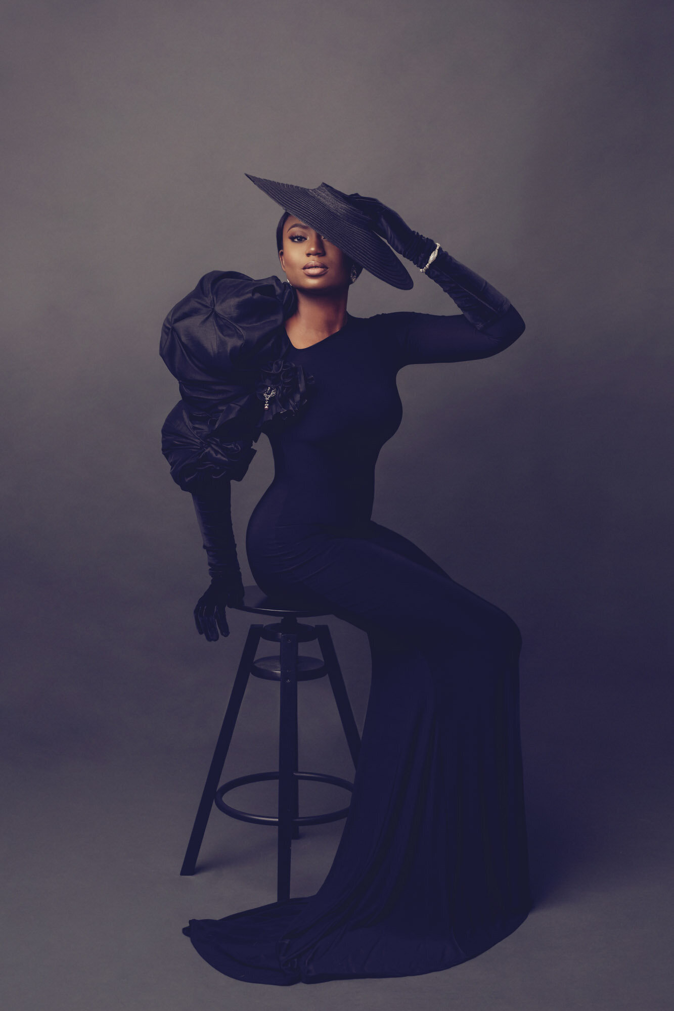Luxury portrait of a woman in a black gown, sitting on a black stool, looking camera center. Houston, TX.