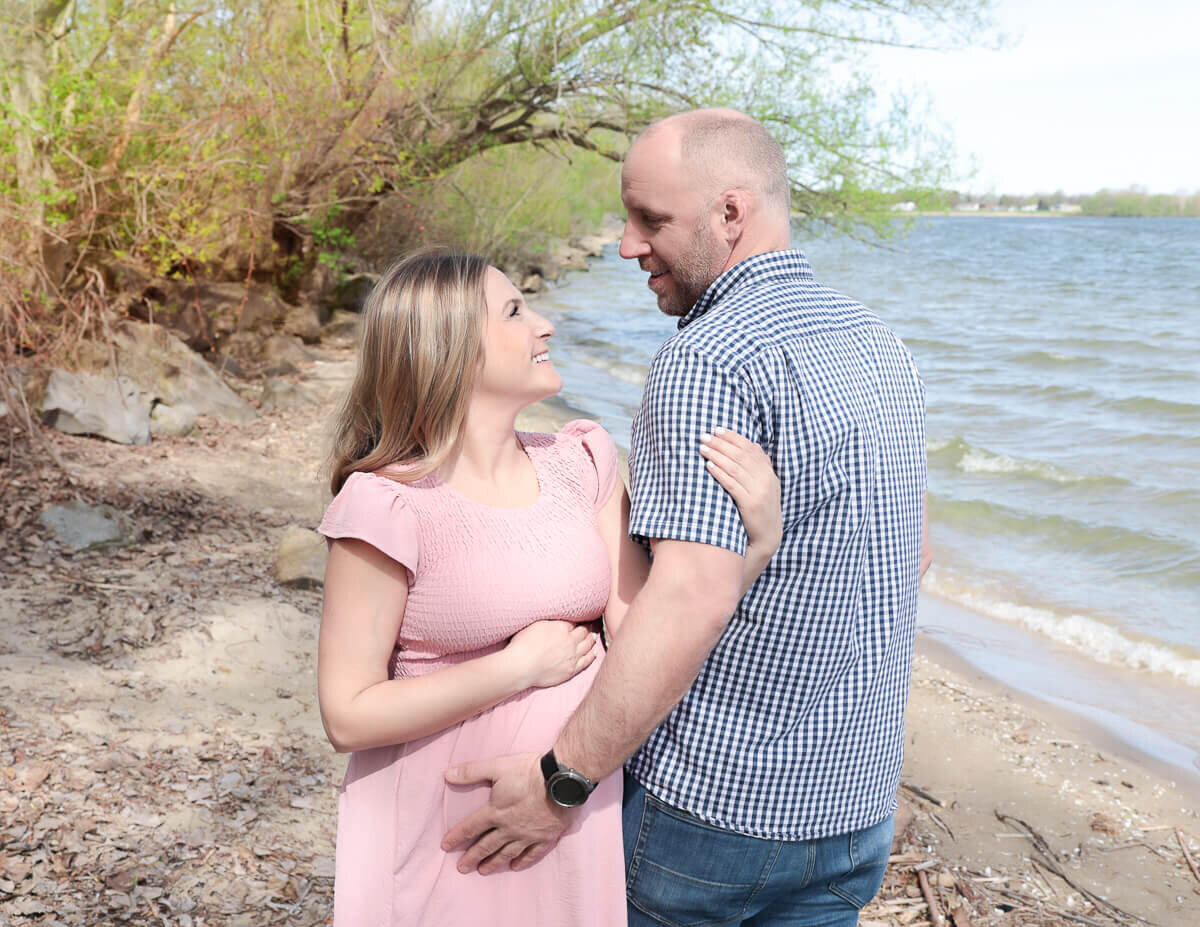 Outdoor maternity photography in Rochester, NY, featuring a loving couple gazing into each other's eyes while gently cradling a baby bump.