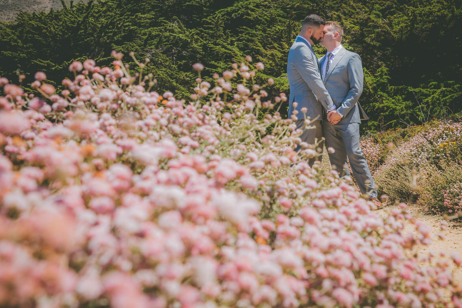 A gay couple kisses with pink flowers in the foreground.