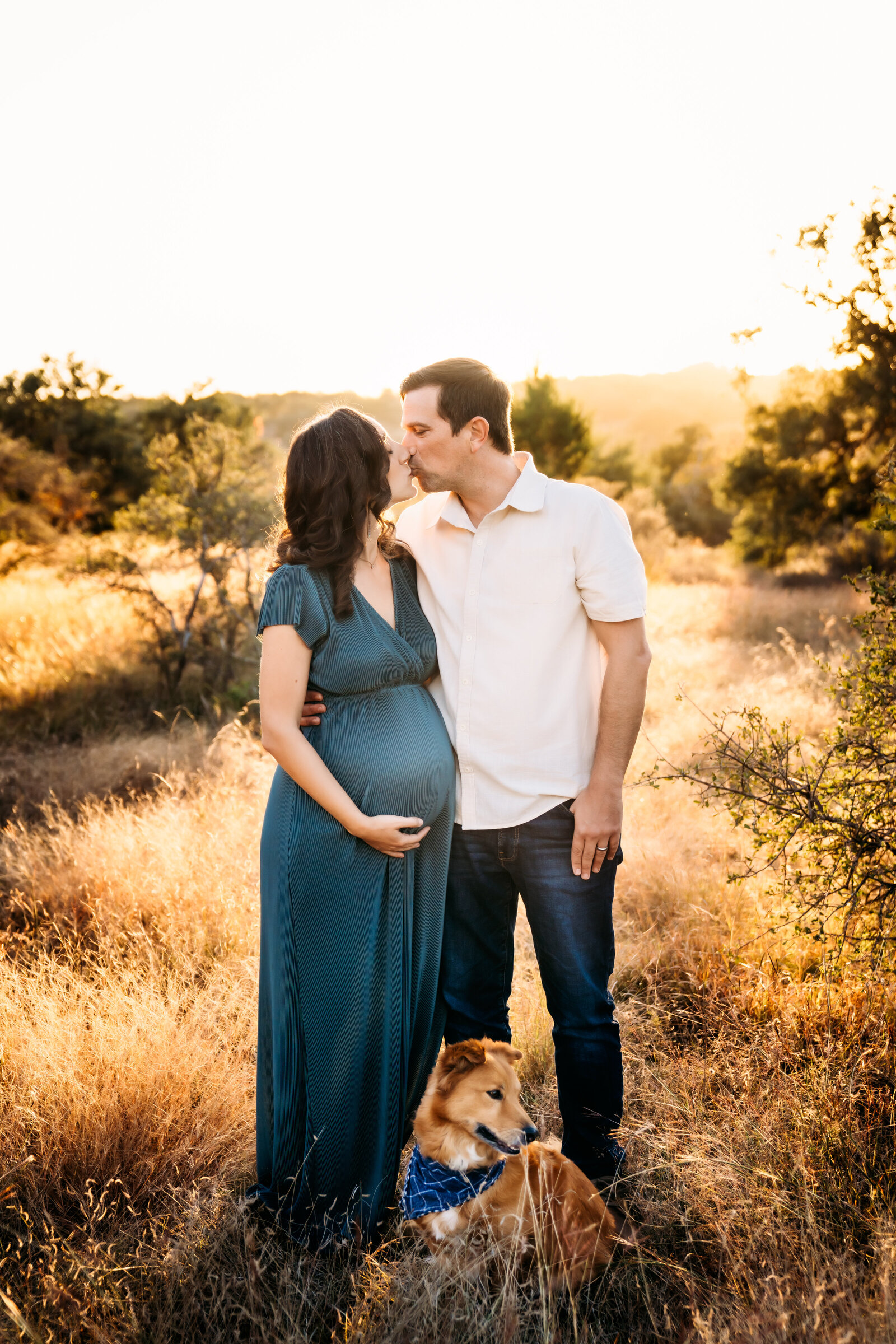 Maternity Photographer, a husband kisses his wife as she holds her pregnant belly, they are in a dry meadow, their dog beside them
