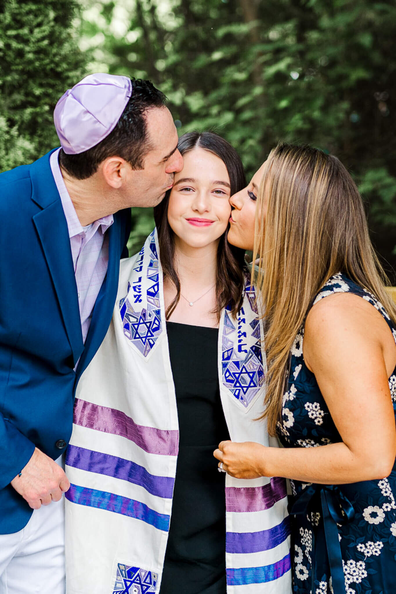 A teen girl in a decorated Tallit is kissed by mom and dad on each cheek while standing outside in a garden