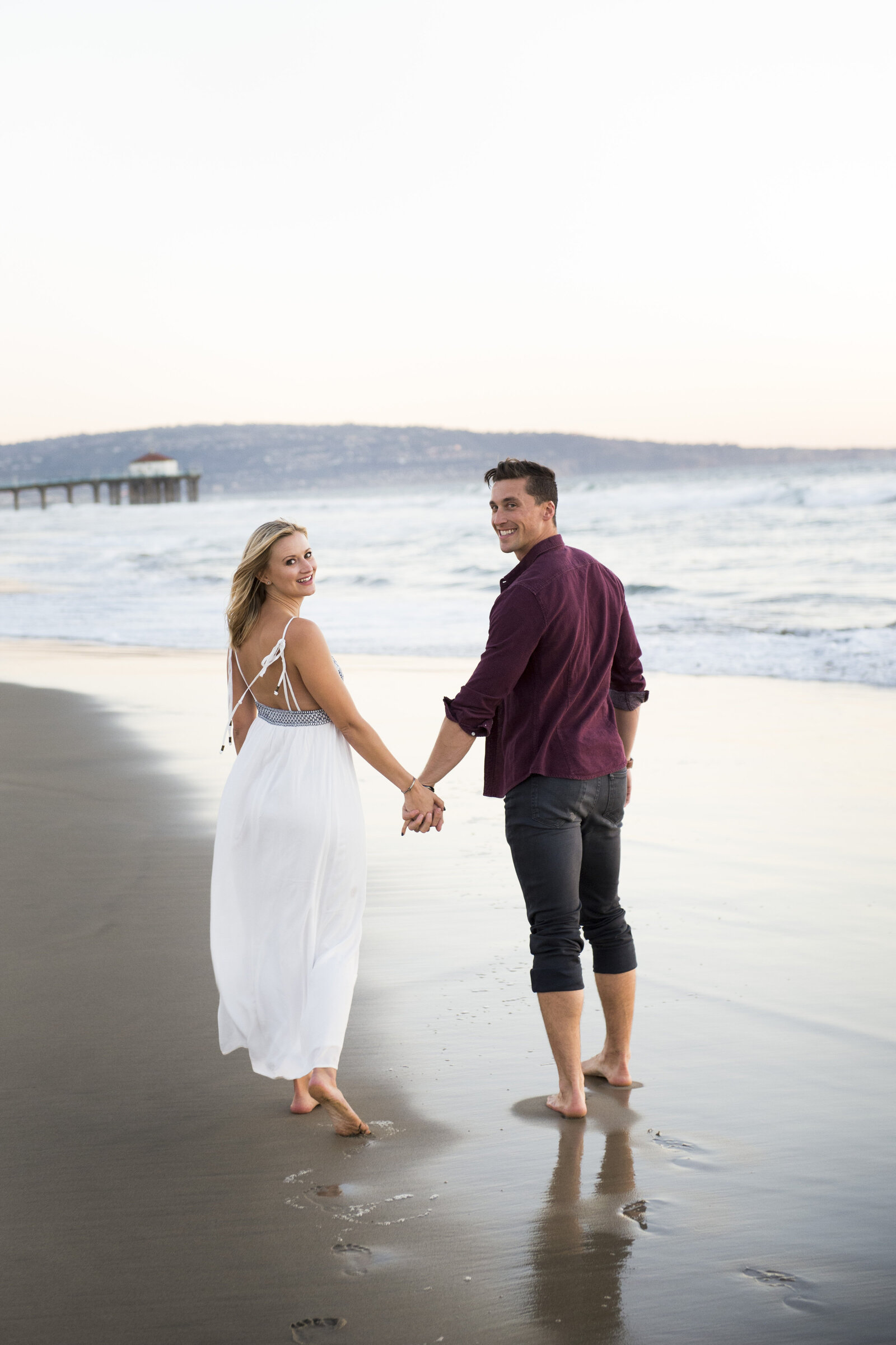 A couple walking down the beach looking back at the photographer during their engagement session.