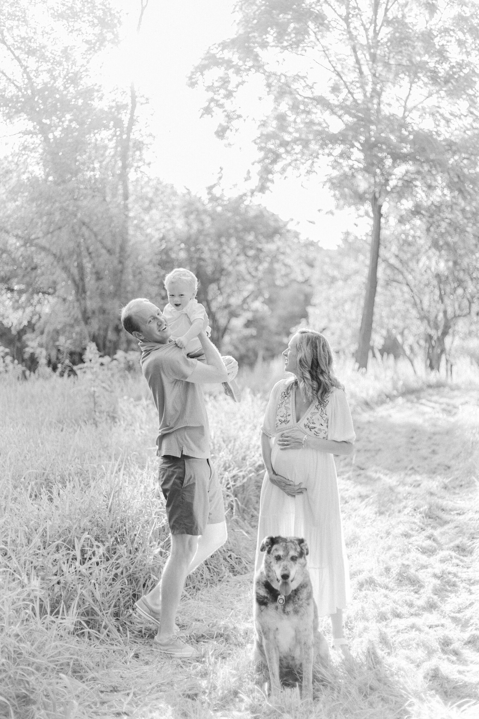 A black and white image of a pregnant mother holding her belly while standing behind their family dog and the father is holding up their toddler boy