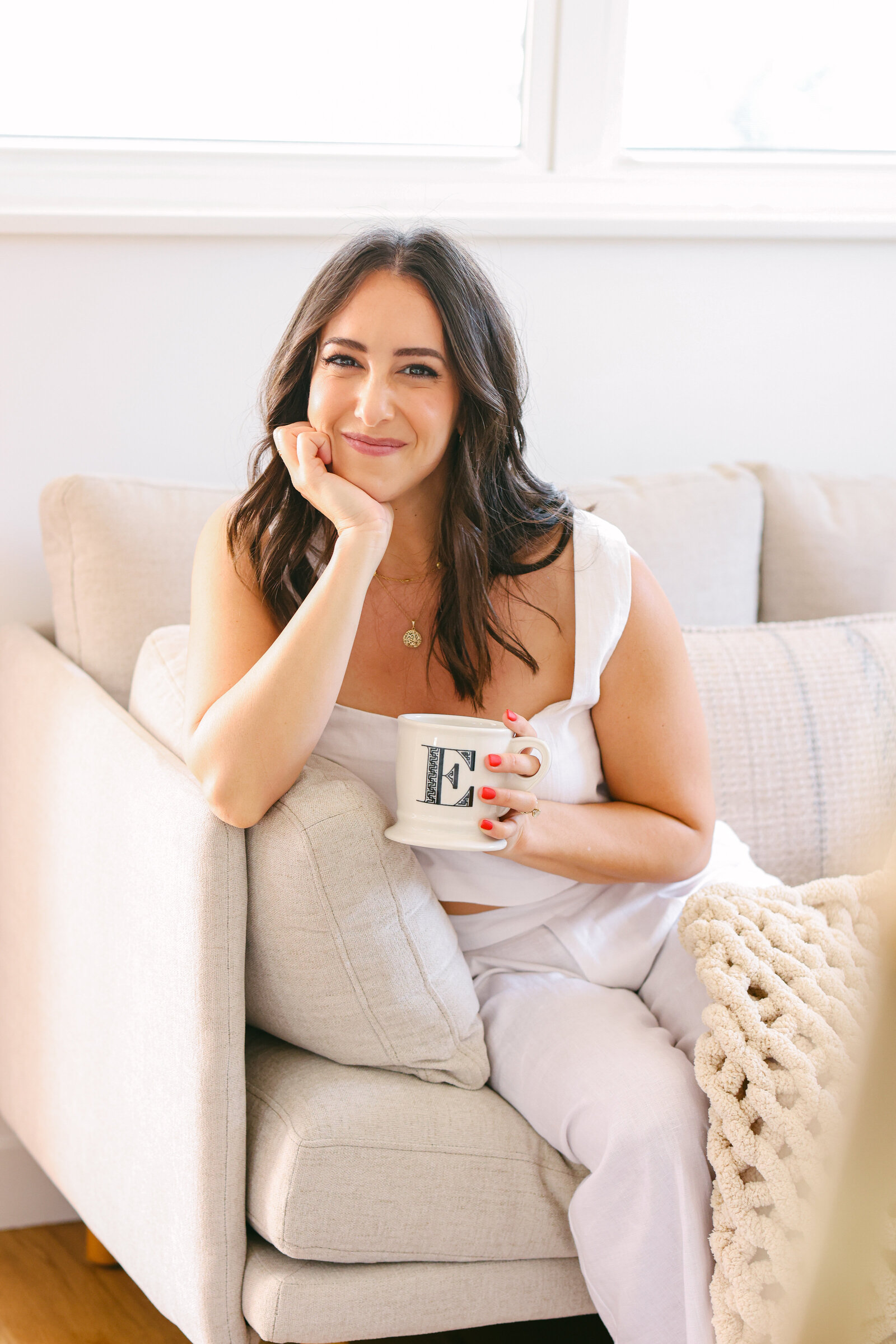 Women Dating Coach Therapist and Life Coach sitting on couch in minimal clean creamy white outfit drinking coffee brand photography session