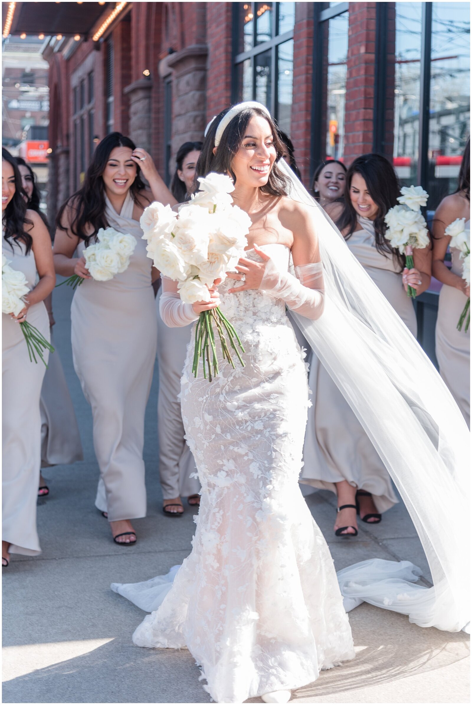 Bride with her bridesmaids and white roses bouquets on Toronto streets - The bride wears a headband with her long hair down matching with her sheer detachable sleeves and sheath silhouette dress with floral bridal lace