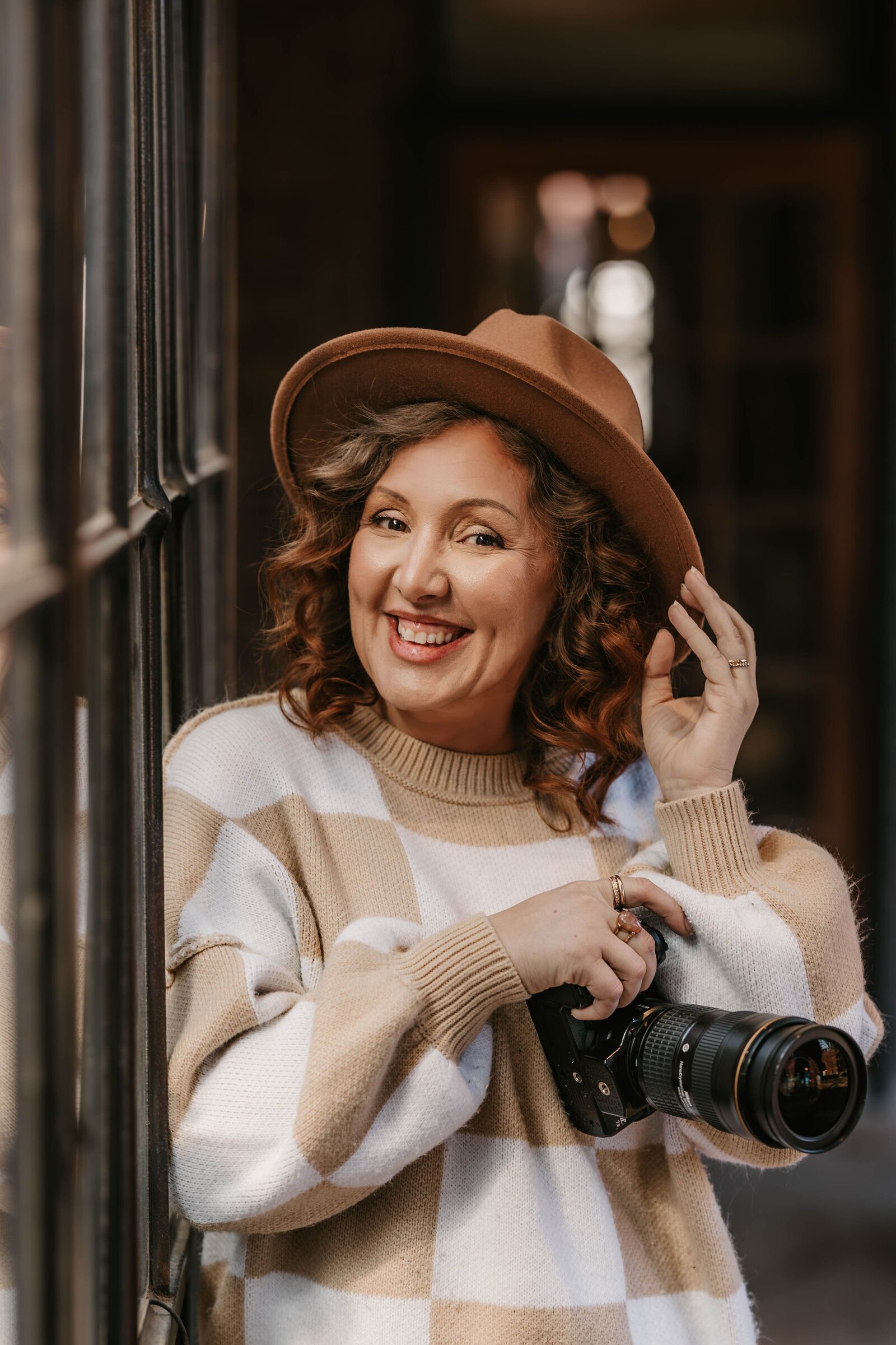 Smiling woman in a hat holding a camera, leaning against a window in cozy, warm lighting—captured behind the scenes at a creative photography retreat.