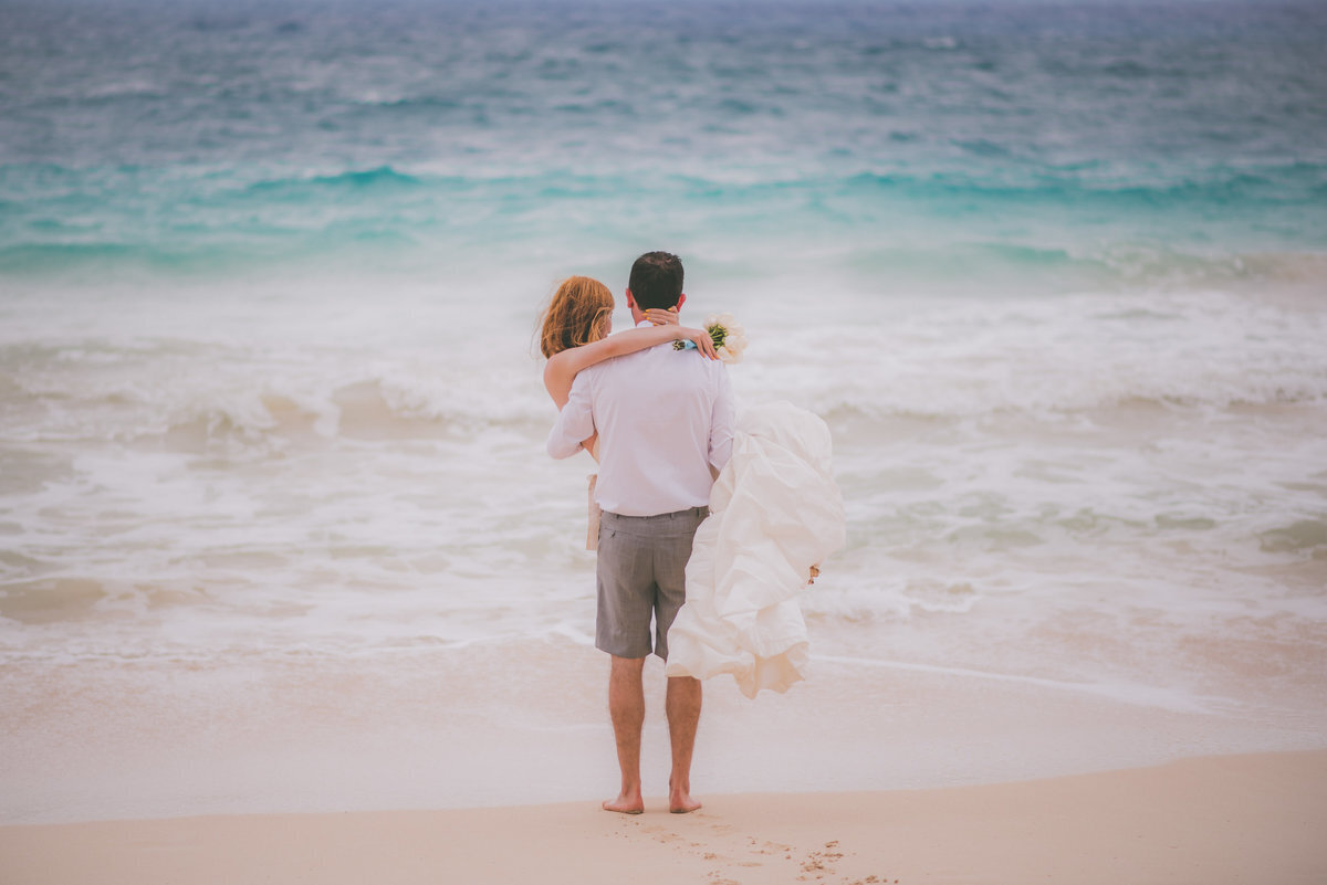 Groom holds bride while they look at the ocean in Hawaii.
