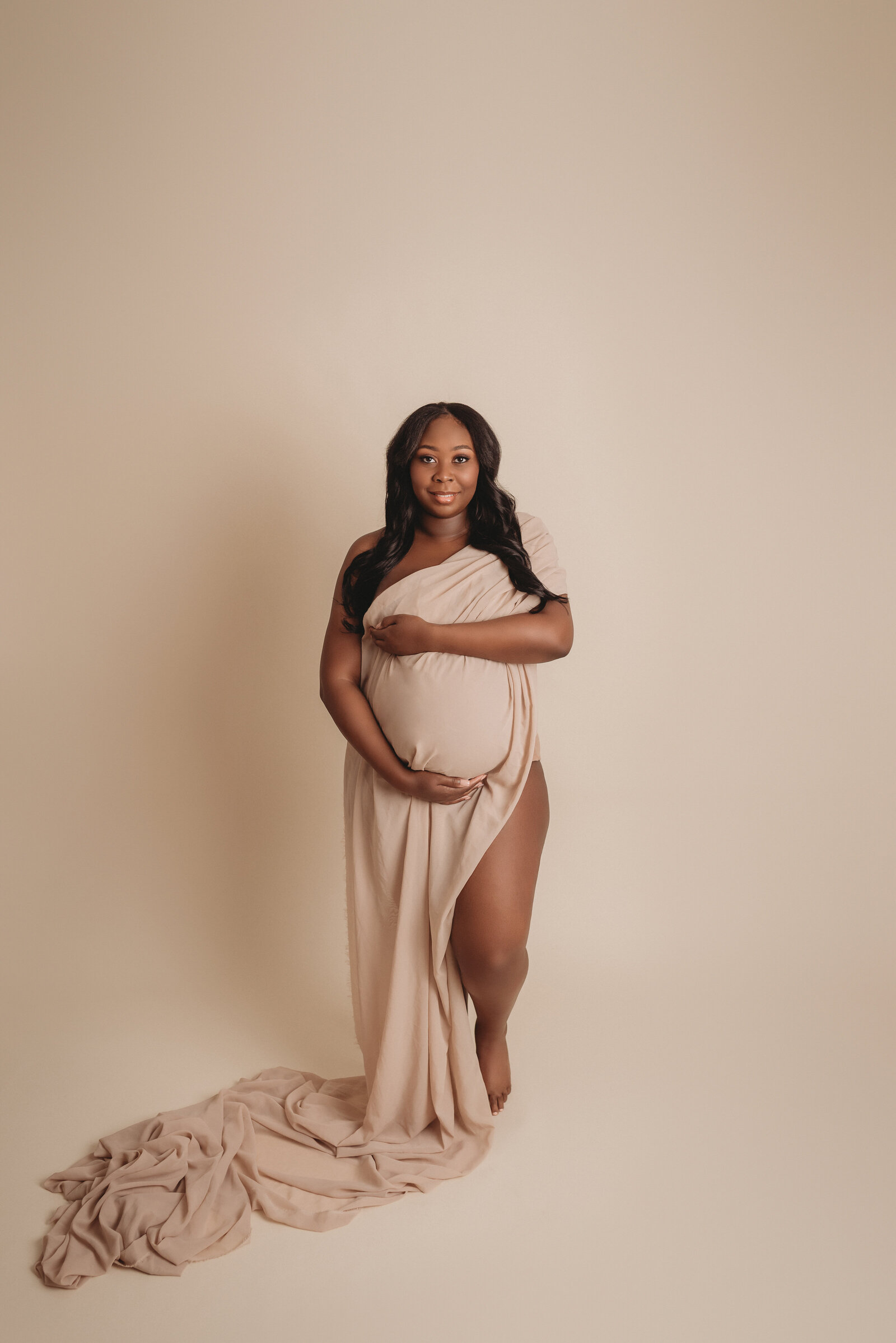 Pregnant woman photographed by atlanta maternity photographer Casey McMinn Photography standing on gray backdrop draped in cream chiffon fabric holding baby bump looking at camera
