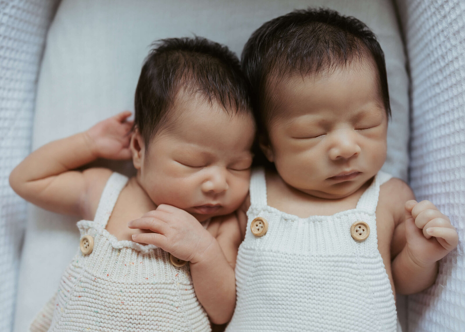 Twin babies sleeping in matching outfits in a white bassinette for their newborn photoshoot.