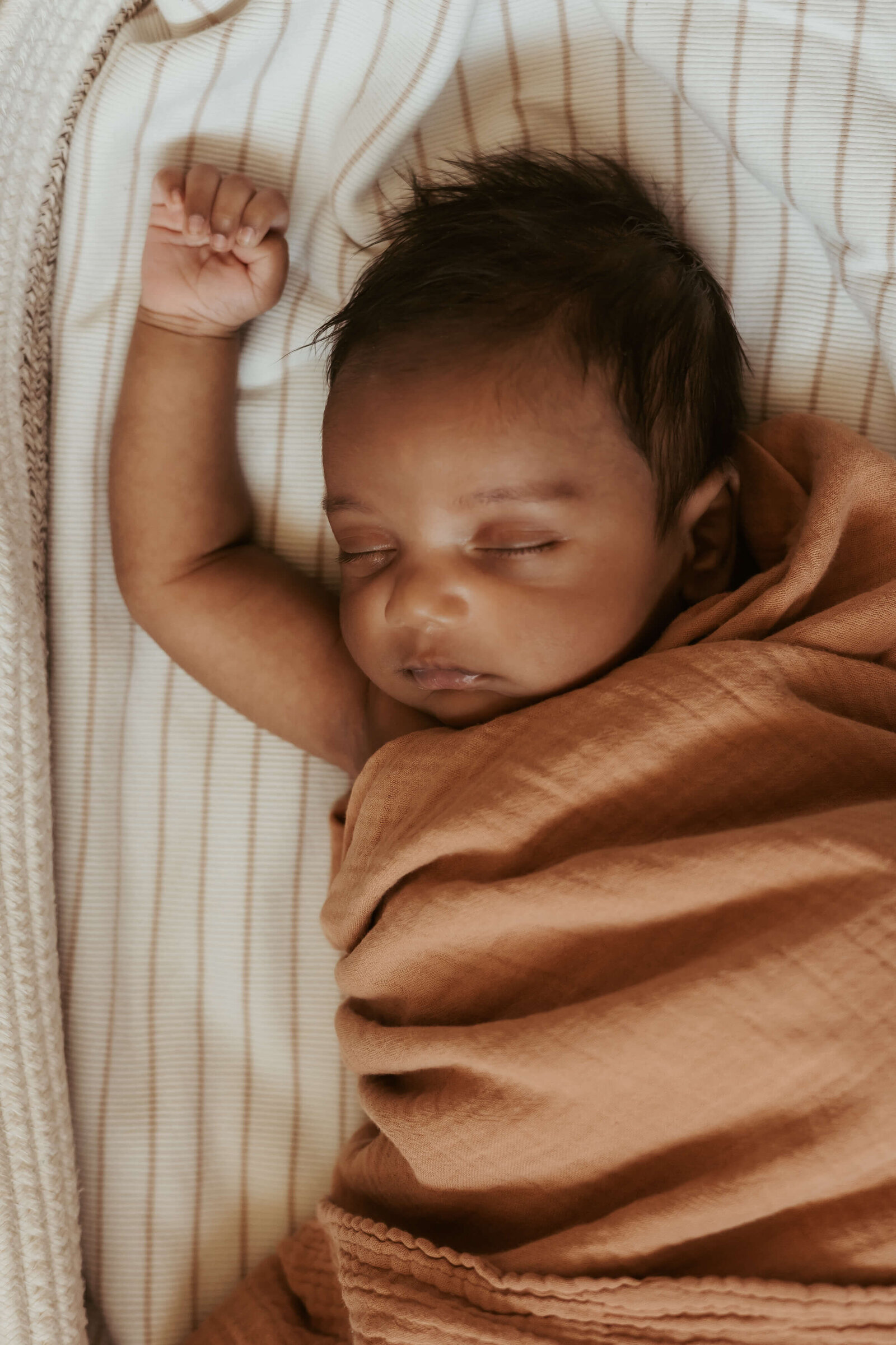 A newborn baby in a brown wrap sleeping with one hand up in his basket.