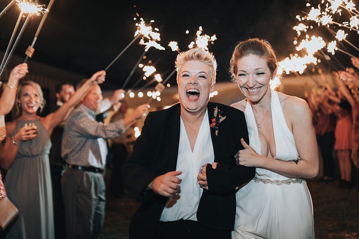 A bride in a black suit and white blouse and a bride in a white jumpsuit with rhinestone belt exit their reception to a sparkler send off