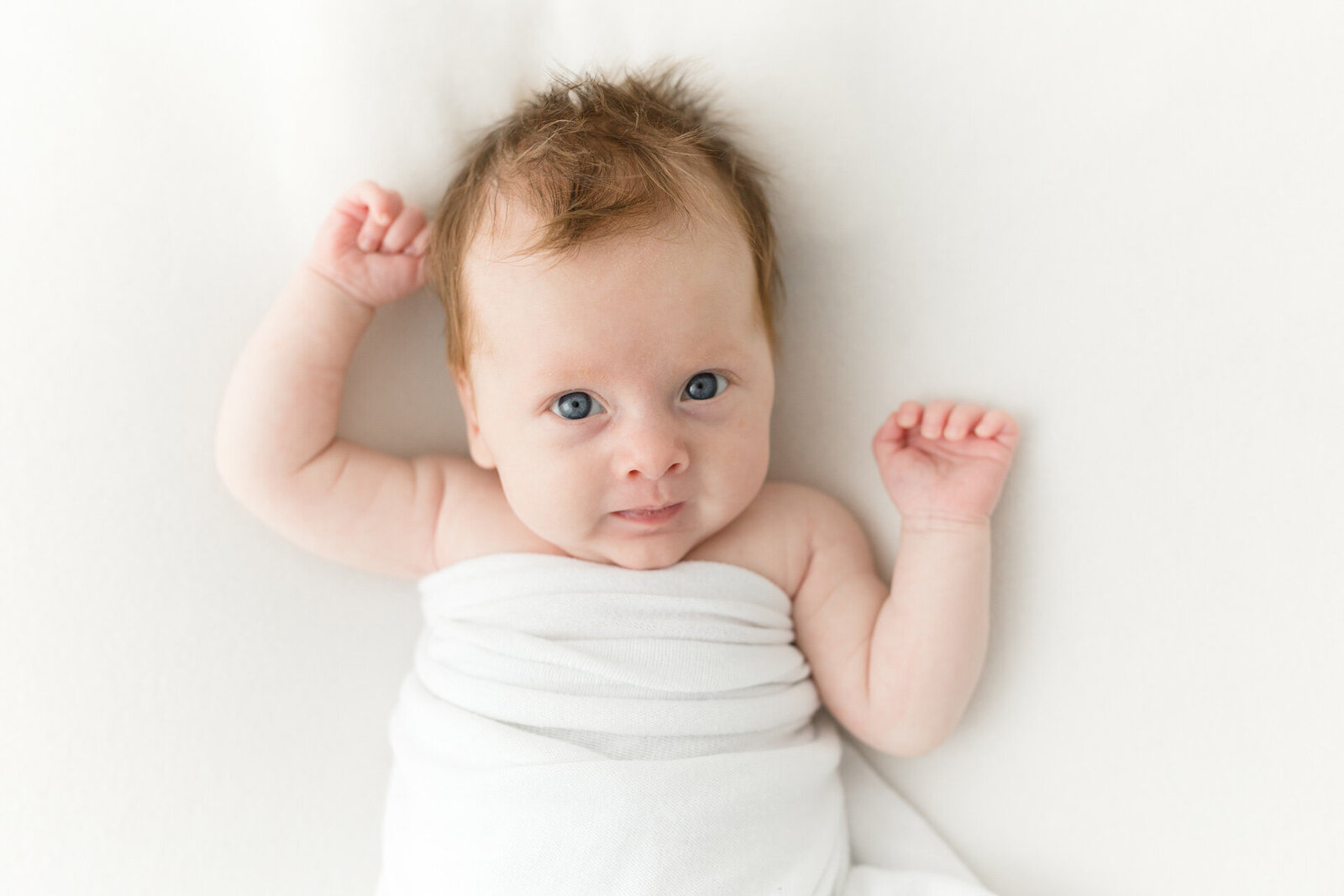 Puristische Neugeborenenfotografie: Baby mit offenen Augen und ausgestreckten Armen  in einer weißen Decke-