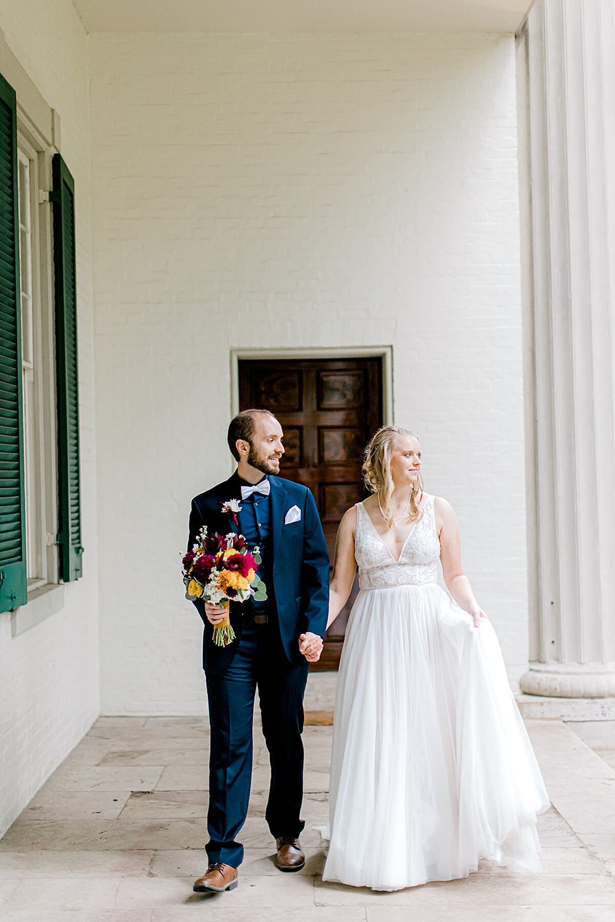 The groom, wearing a navy suit, holds the bride's brightly colored bouquet as she leads him by the hand.. The bride is wearing a beauiful satin aline gown with rhinestone accents.