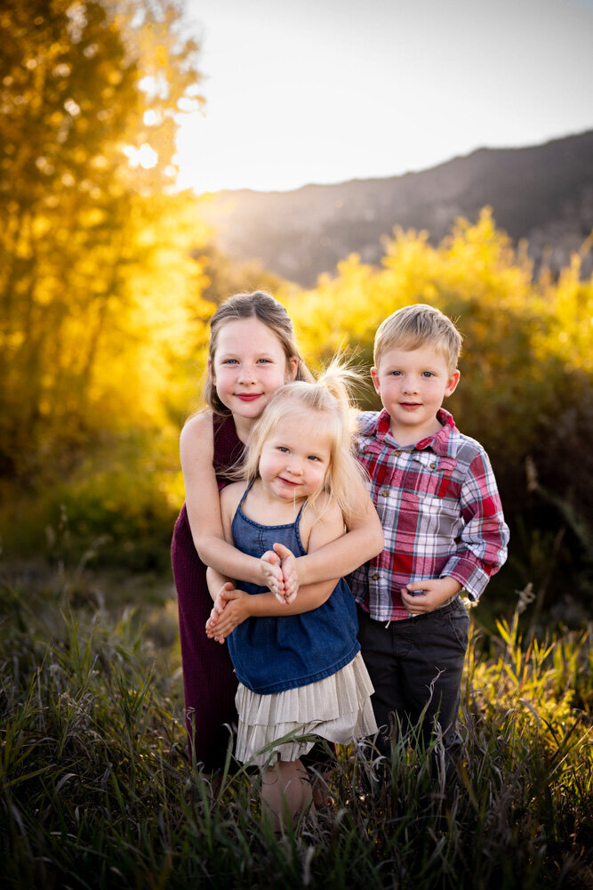 Three children stand in front of fall foliage and hug each other