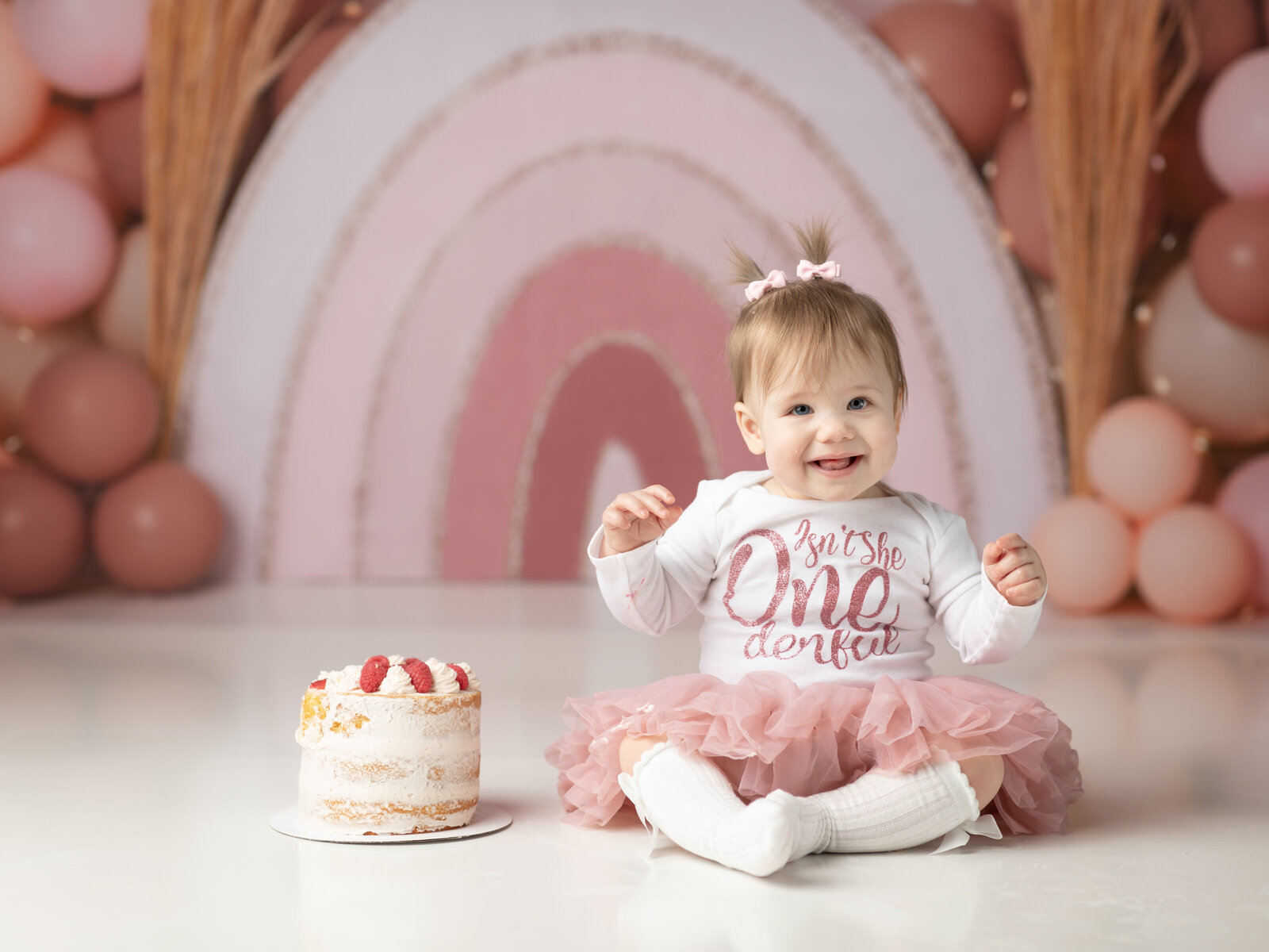 one year old girl sitting with cake and boho rainbow backdrop for cake smash photoshoot