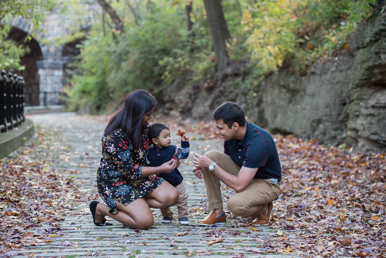 family kneels on cobblestone pathway surrounded by fall leaves in schenley park in pittsburgh's oakland neighborhood