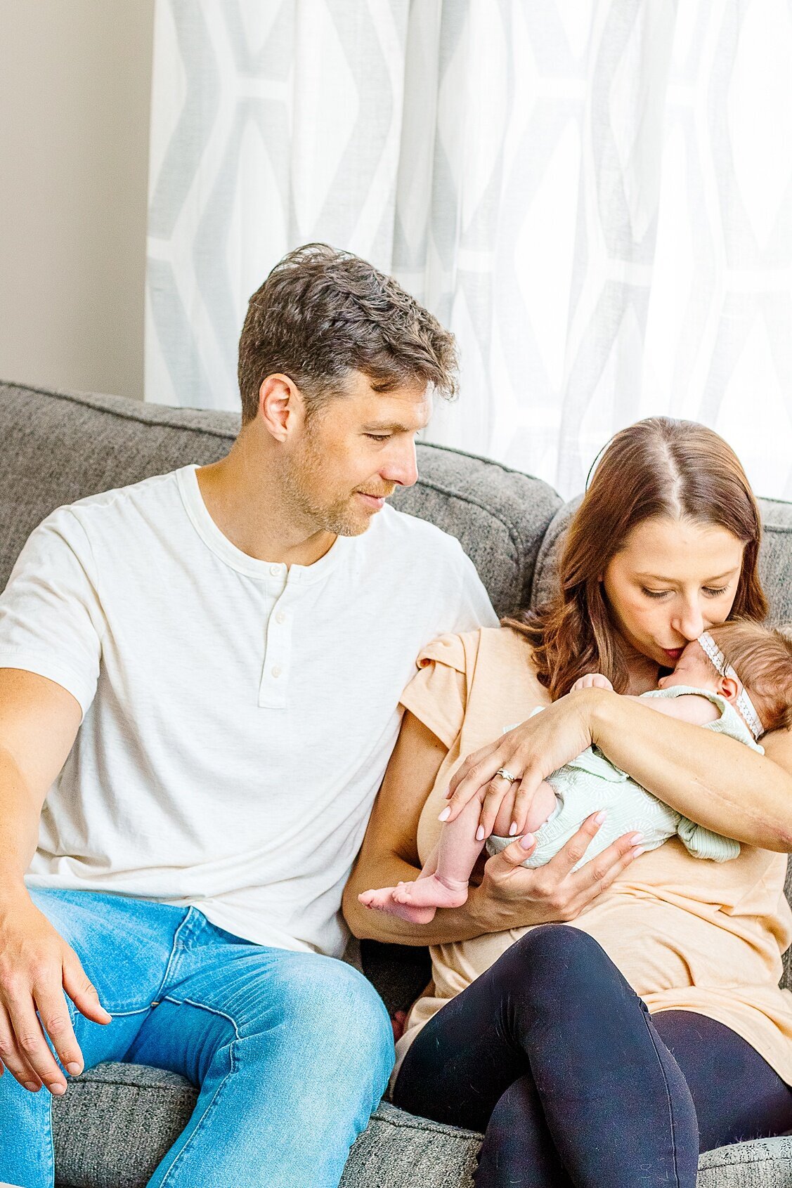 Mom is holding her baby girl while dad looks on with a smile.  mom is kissing the top of baby girls face during a Lifestyle Newborn Session in Wexford PA