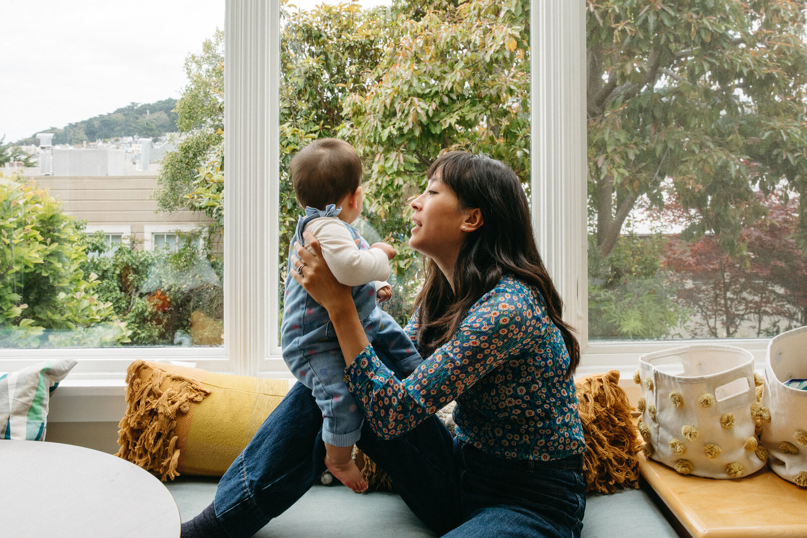 mom in floral top and wide leg jeans sitting in window seat holding baby daughter