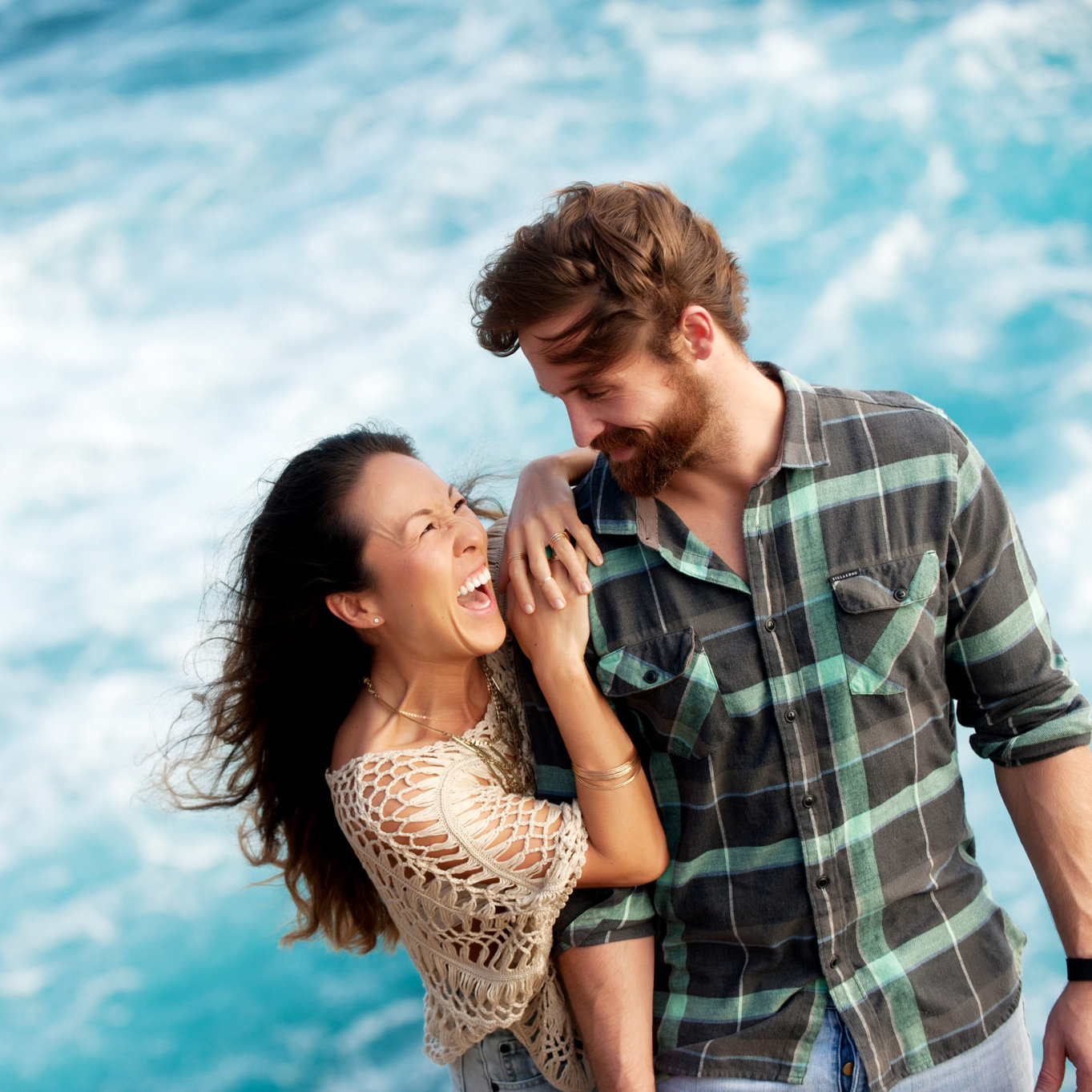 an attractive couple looks and laughs at each other standing above bright blue waters at spitting caves in hawaii
