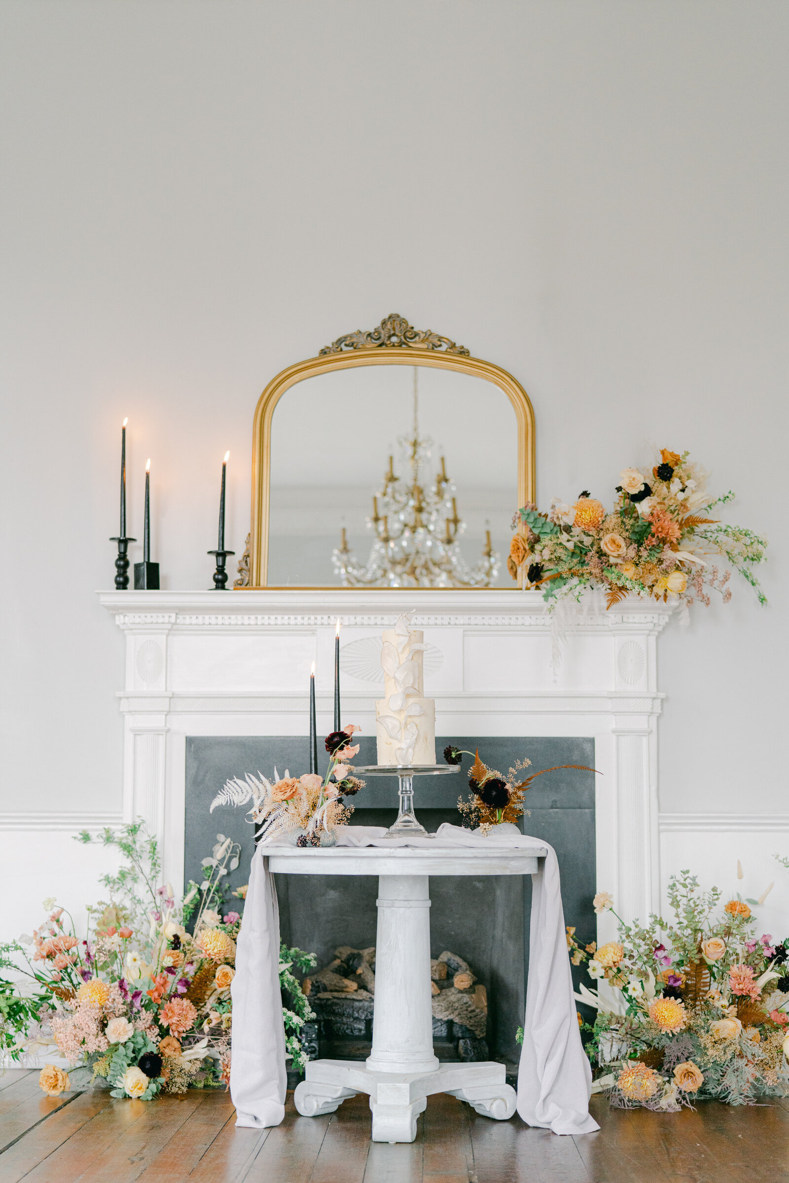 wedding cake on a stand in front of mantel with floral arrangements all around