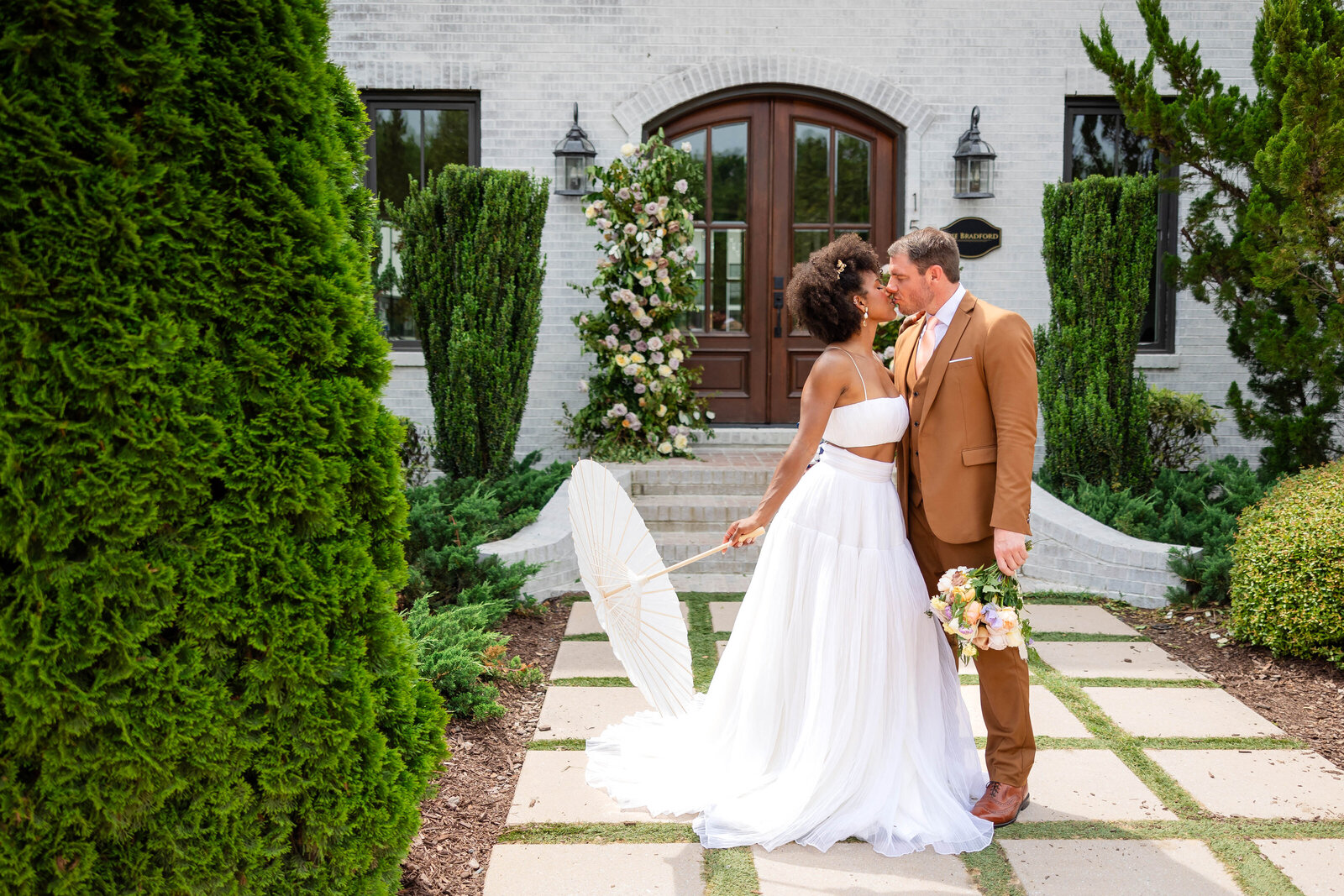 Bride and groom standing outside of a large white  luxury barn type huilding with black and brown edging and wooden accents.  Bride in white with applique flowers on the bodice of the fitted and flared gown.  Wearing gold flower earrings.  Groom in a black tux with white shirt and black bow tie.  Both holding their vows and reading to each other before seeing each other.  Groom on one side of the building and Bride on the other with just the corner of the building between them.  They are holding hands.