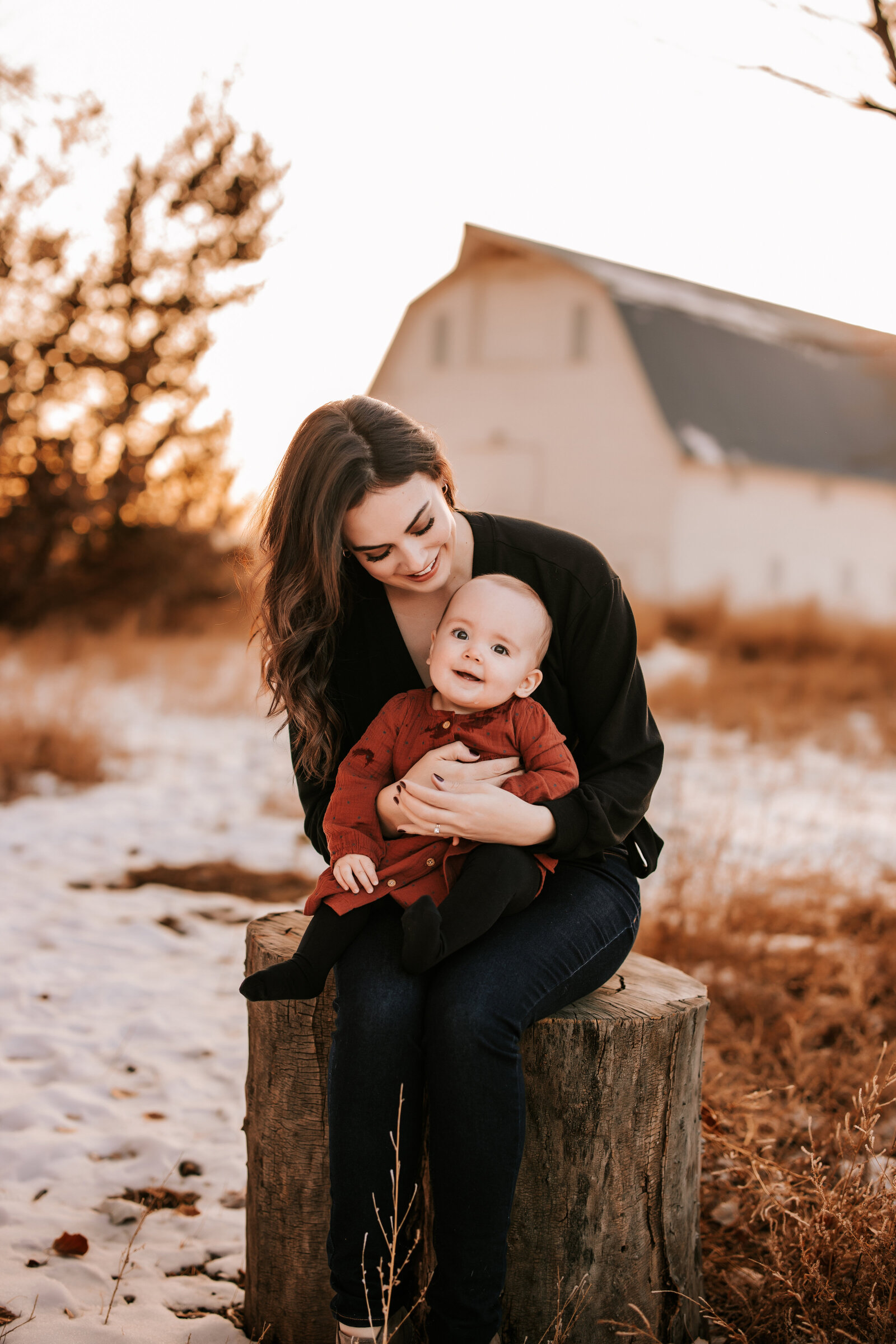 mom and baby girl sit on tree stump in front of a white barn with snow on the ground