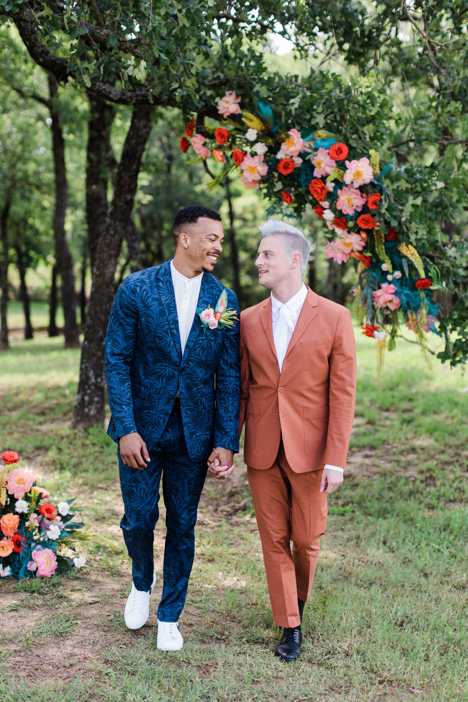 Two grooms walking together while holding hands and looking into each other's eyes at Bella Cavalli Events in Aubrey, Texas. They are surrounded by many trees and some large, colorful floral arrangements. The African American groom on the left is wearing a blue, floral suit and boutonniere. The Caucasian groom is on the right and is wearing a reddish orange suit.