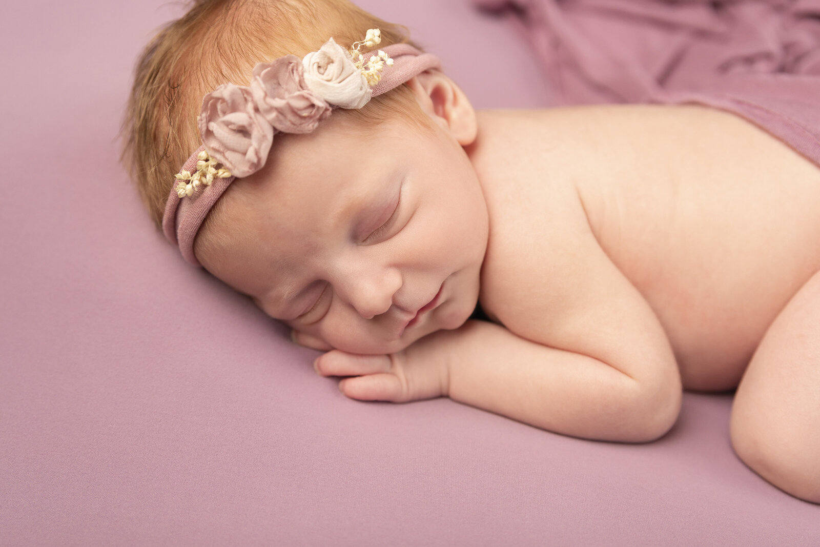 Charlotte  Newborn baby girl portrait wearing a pink headband with flowers laying down sleeping on a matching pink blanket, almost appears to smile, white baby with bright read hair