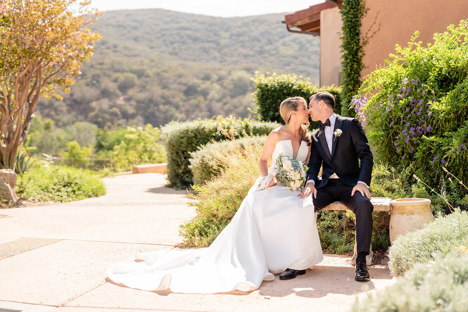 A bride and groom sitting next to each other almost kissing on a bench outside at Vista Valley Country Club.
