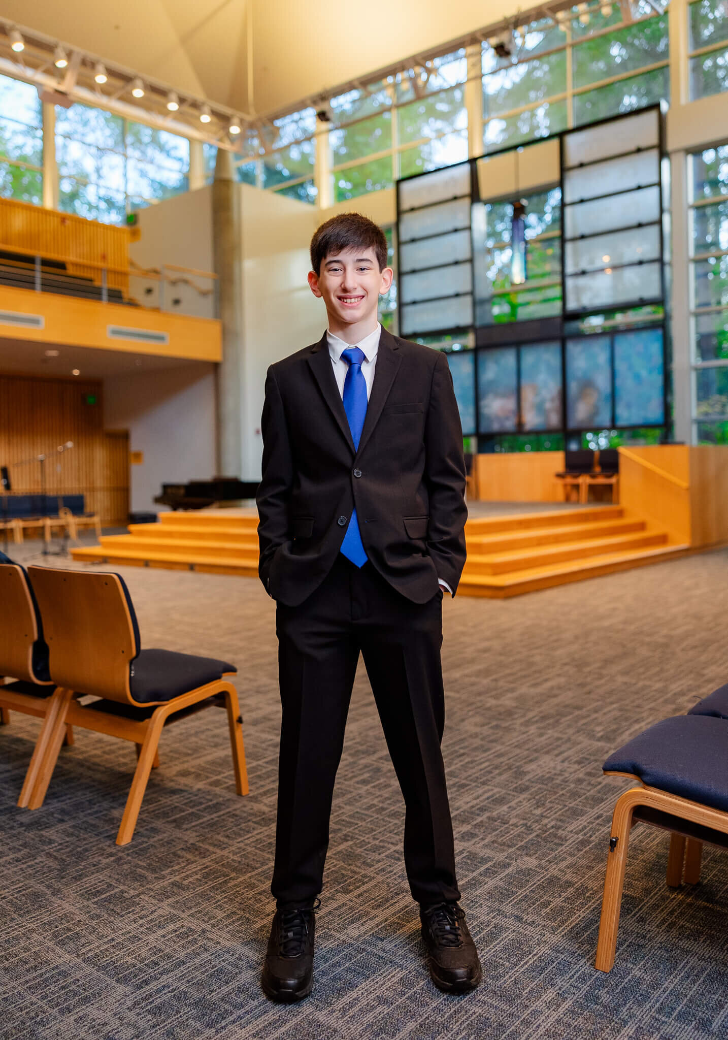 A teen boy in a black suit and blue tie stands in the temple with hands in pockets during a Bellevue Bar and Bat Mitzvah Photography session