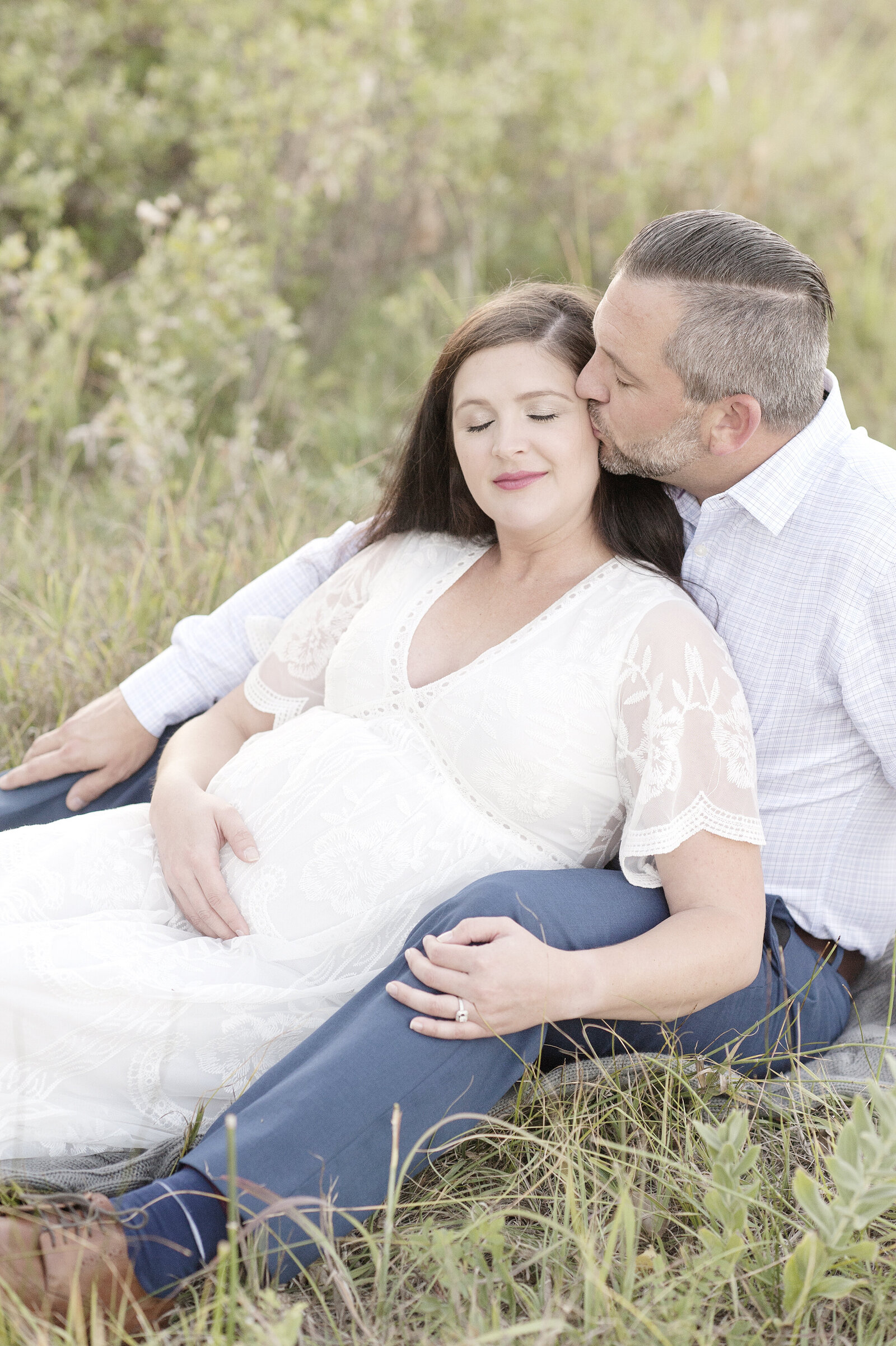 Dad kissing pregnant woman while sitting in grass