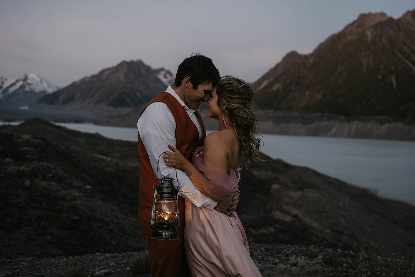 A couple embraces at dusk by a lake, holding a lantern with mountains in the background—capturing true connection, much like moments found at a Utah desert photography retreat focused on real connection and growth.