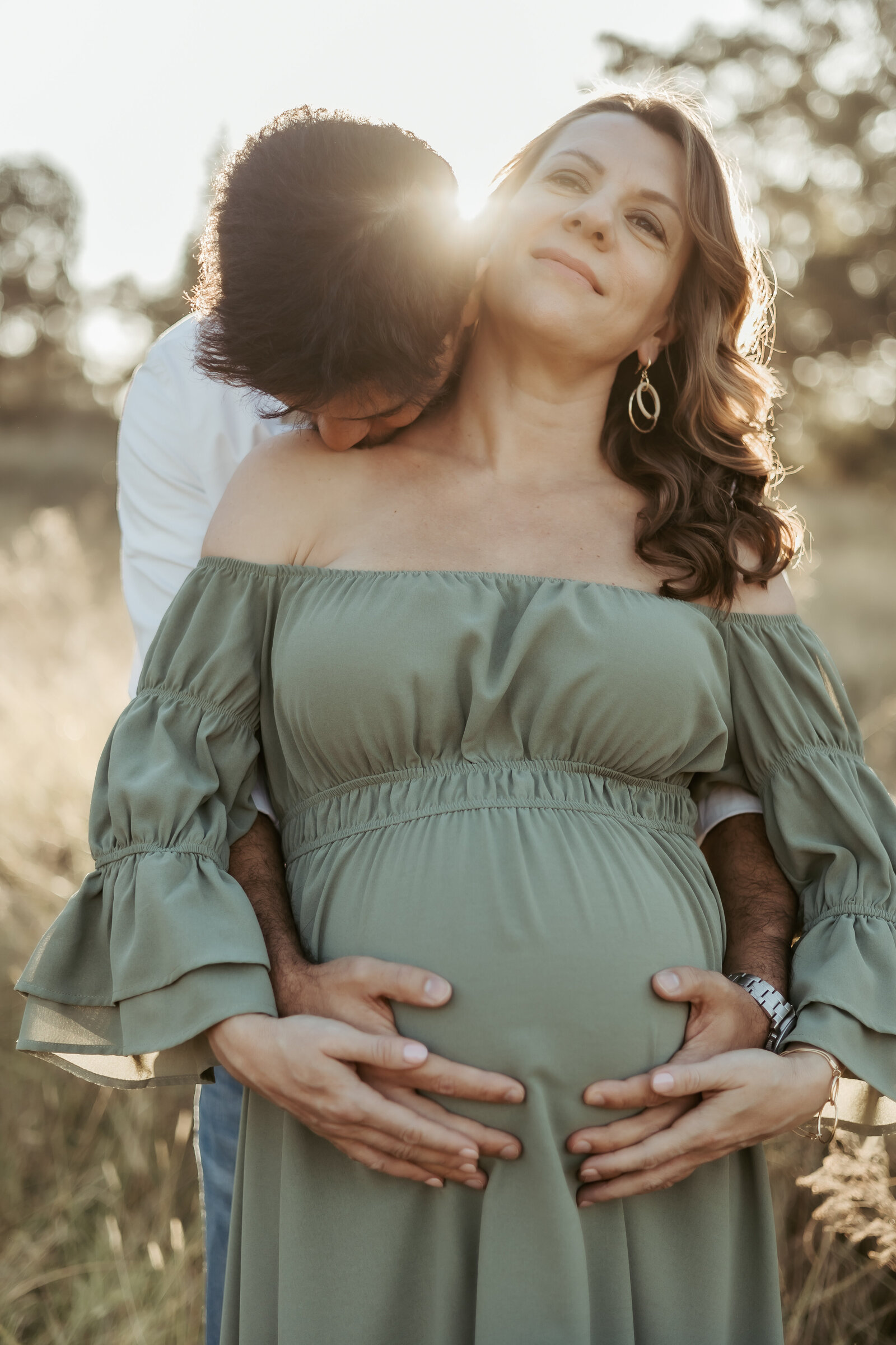 Pregnant mum in a green dress holding her growing bump  while partner is behind her kissing her neck and the sun glows between their heads