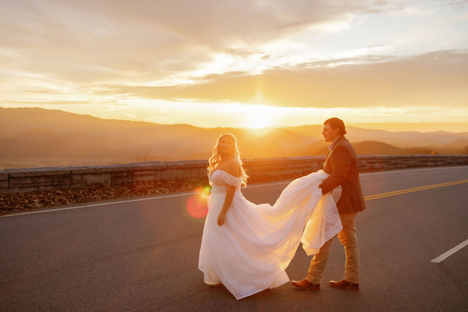 gatlinburg photographers photographs sunset wedding photos with groom holding the brides wedding dress as they walk together across the foothills parkway in the Smoky Mountains 