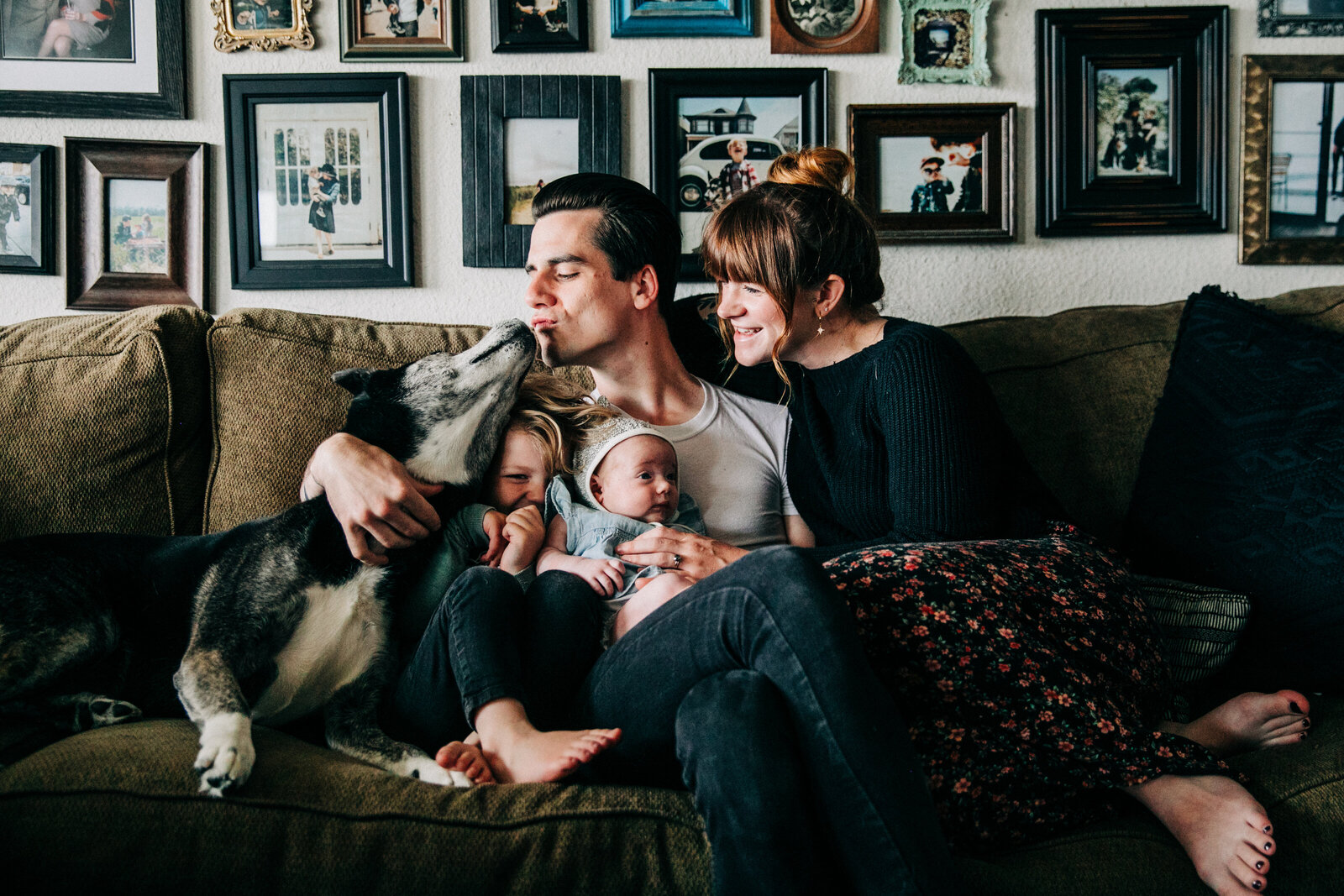 dad kissing dog and sitting with mom on a big couch with newborn baby girl and toddler son with a wall full of picture frames behind them