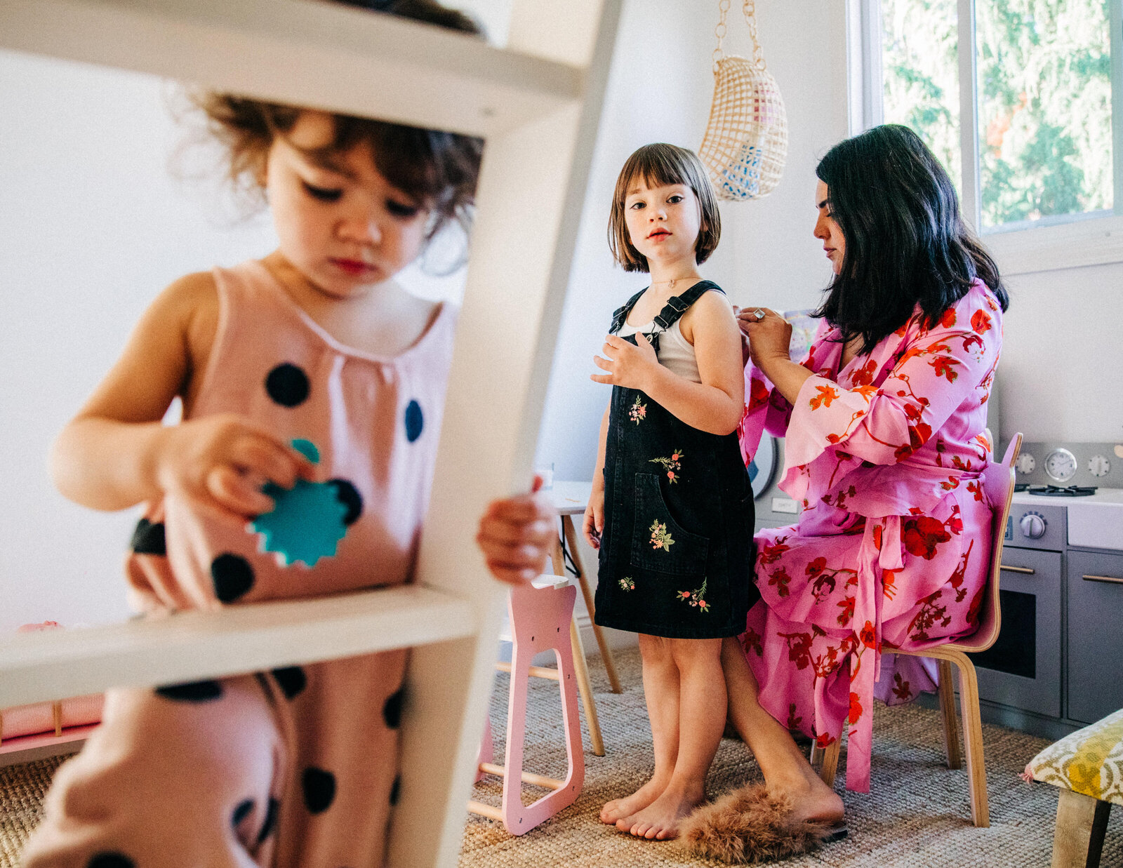 photo of mom in pink dress helping daughter get dressed in black overall dress with younger daughter in the foreground playing on a bunk bed ladder