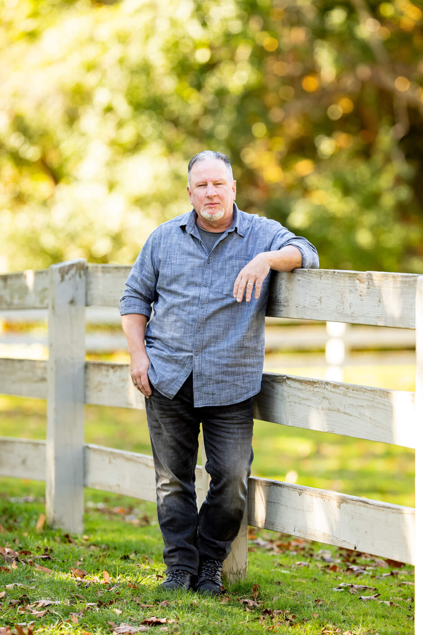 Portrait of an adult male in a blue button down shirt and black jean leaning on a white fence in Beverly MA Endicott Park