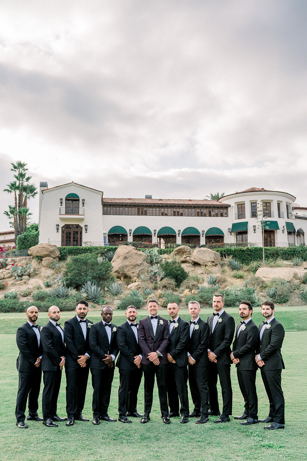 Groomsmen pose in classic black tuxedos in front of the Hummingbird Nest Ranch estate, photographed in timeless editorial style.