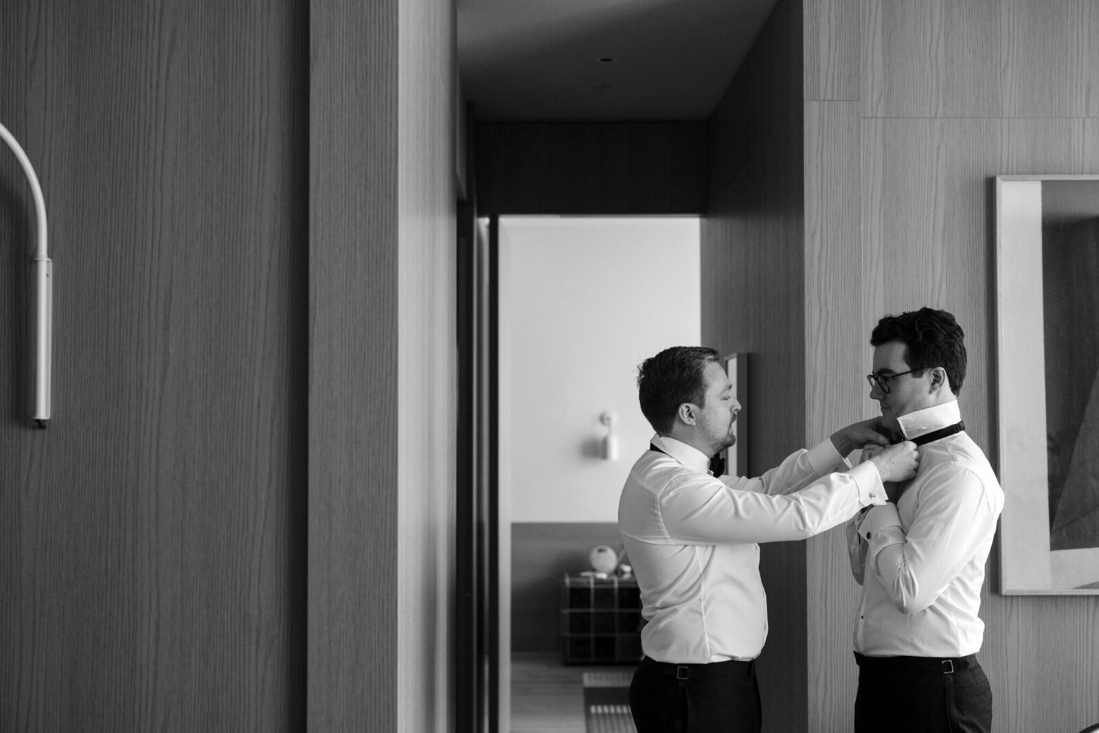A man putting a tie on a groom to prepare for his wedding, in a black and white photo by Ada and Ivy