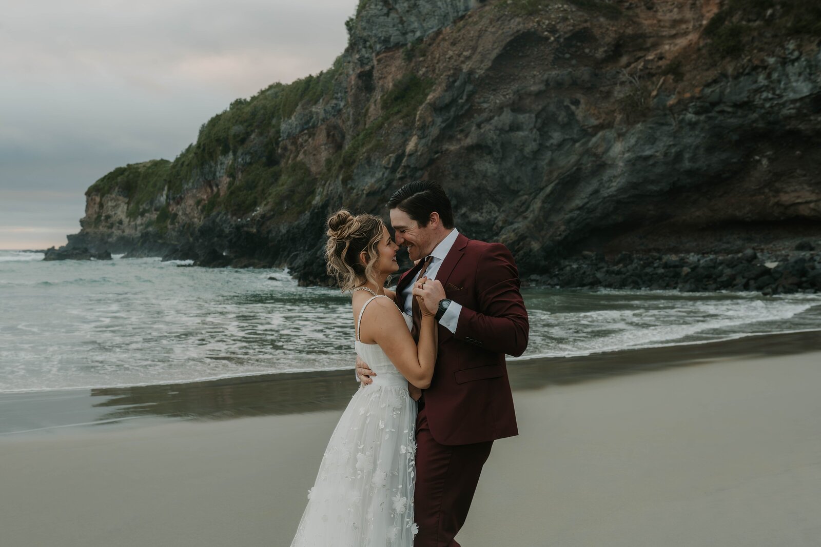 Bride and groom embrace and smile on a beach, with cliffs and ocean waves in the background—a moment reminiscent of a California photography retreat for storytellers and soul seekers.