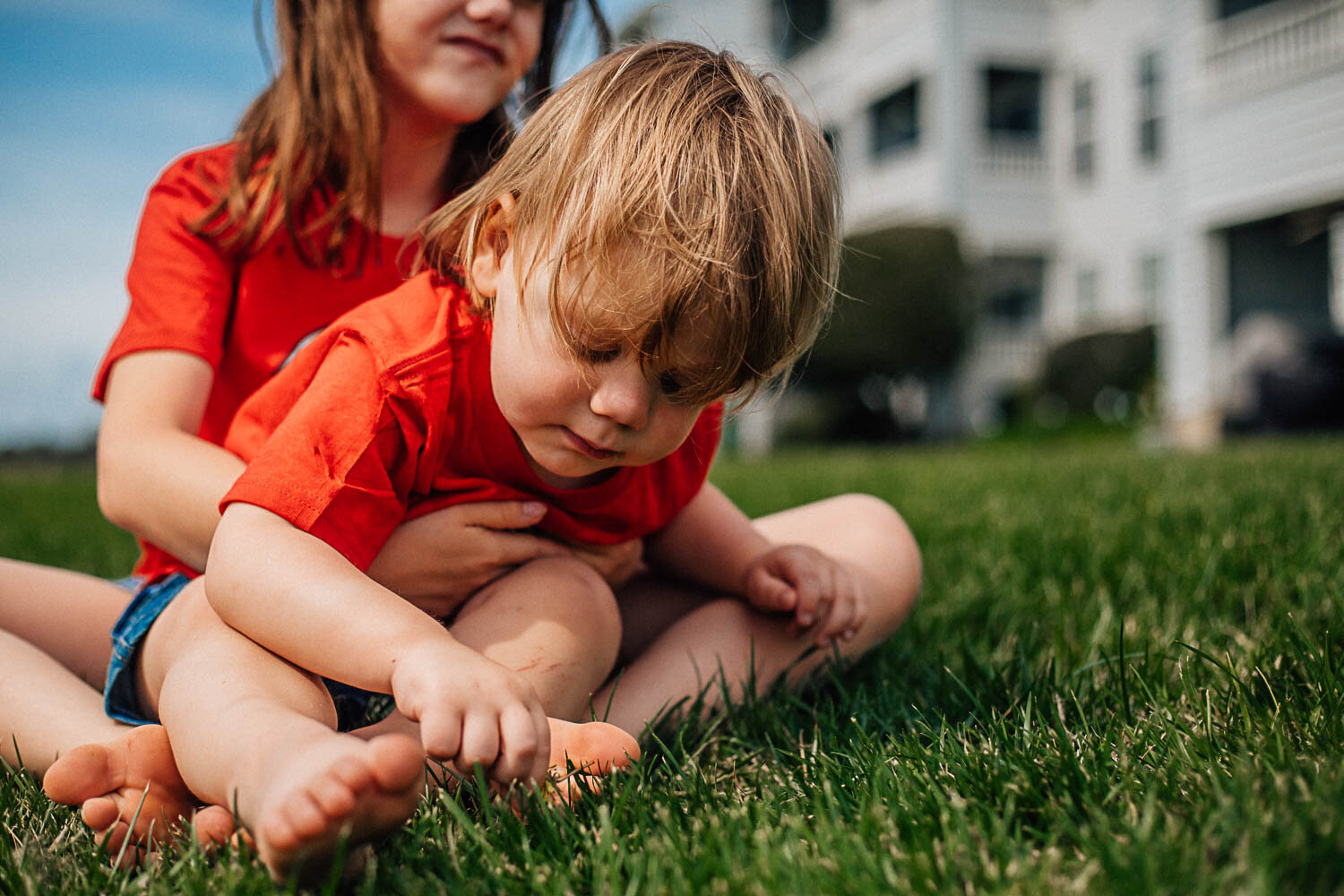 Hampton Roads family photographers capture little girl holding her toddler sibling as they both sit together in the grass for family photos in Hampton Roads