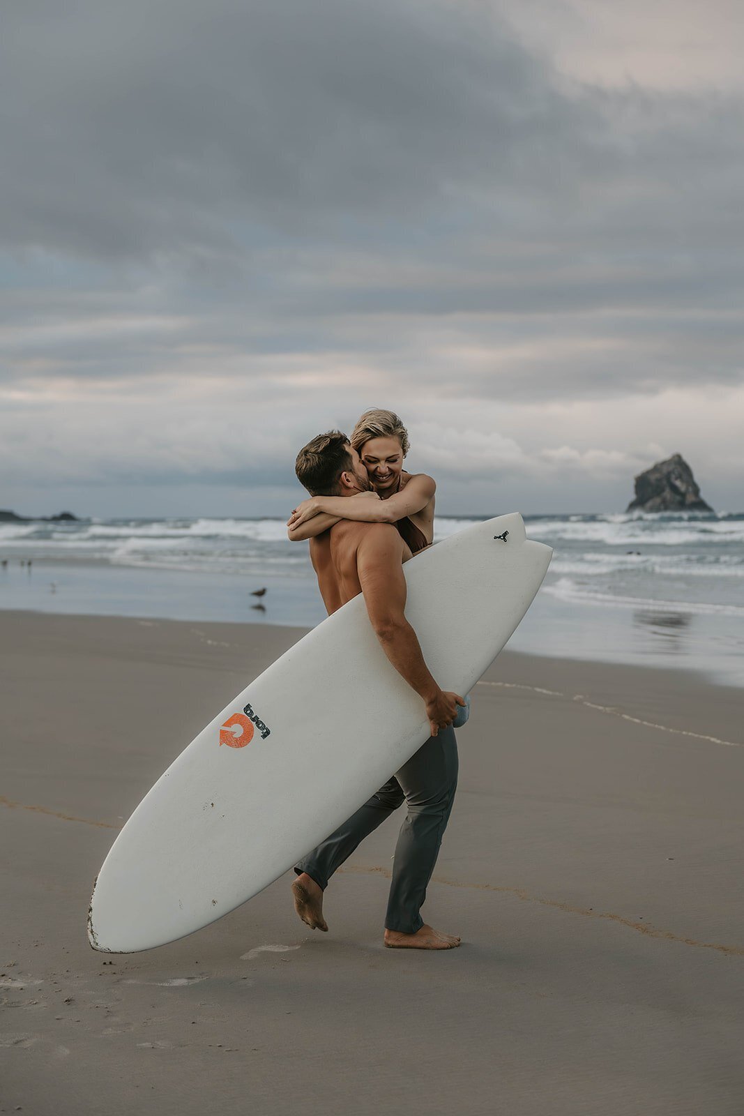 A man carries a woman and a surfboard on the beach, both smiling with waves in the background—capturing the spirit of an Oregon coast photography retreat for wild-hearted artists.