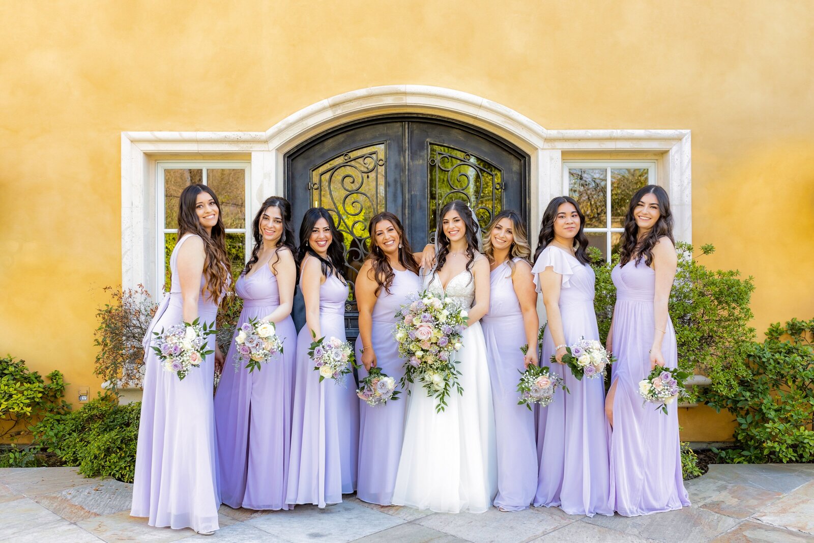 Bride stands in the center with bridesmaids who are dressed in different shades of purple smiling and looking at camera at Arden Hills in Sacramento, captured by Philippe Studio Pro.