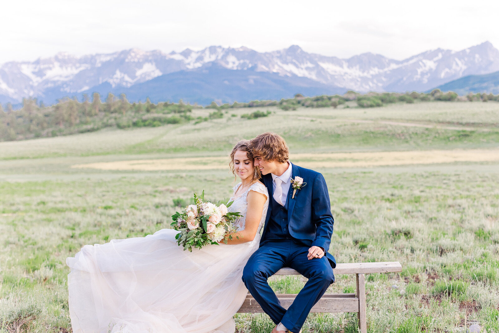 groom kisses bride on bench in front of the mountains