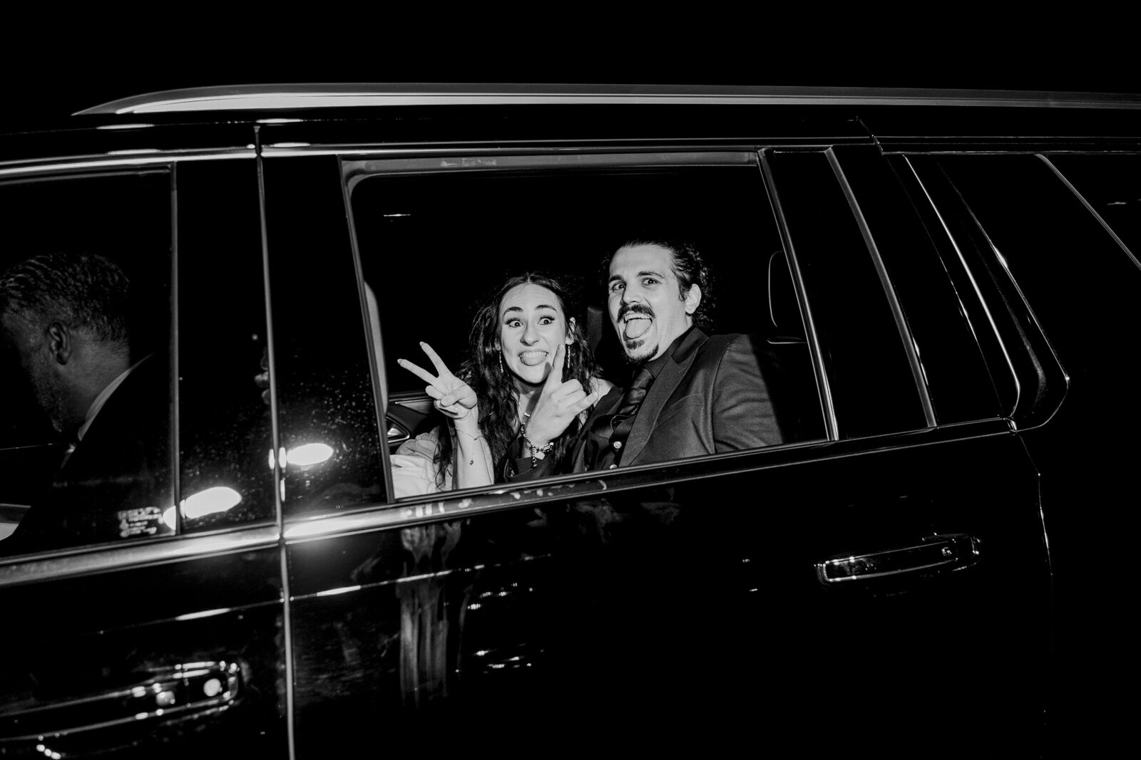 A black and white candid photo of a bride and groom playfully saying goodbye to their guests in their exit car after their wedding reception at The Lodge at The Springs in Denton, Texas. The bride and is on the left and is holding up a peace sign with one hand and has a large surprised, playful smile. The groom is on the right and is holding up the rock and roll hand sign and is sticking his tongue out. A drive can be seen in the front seat of the car.