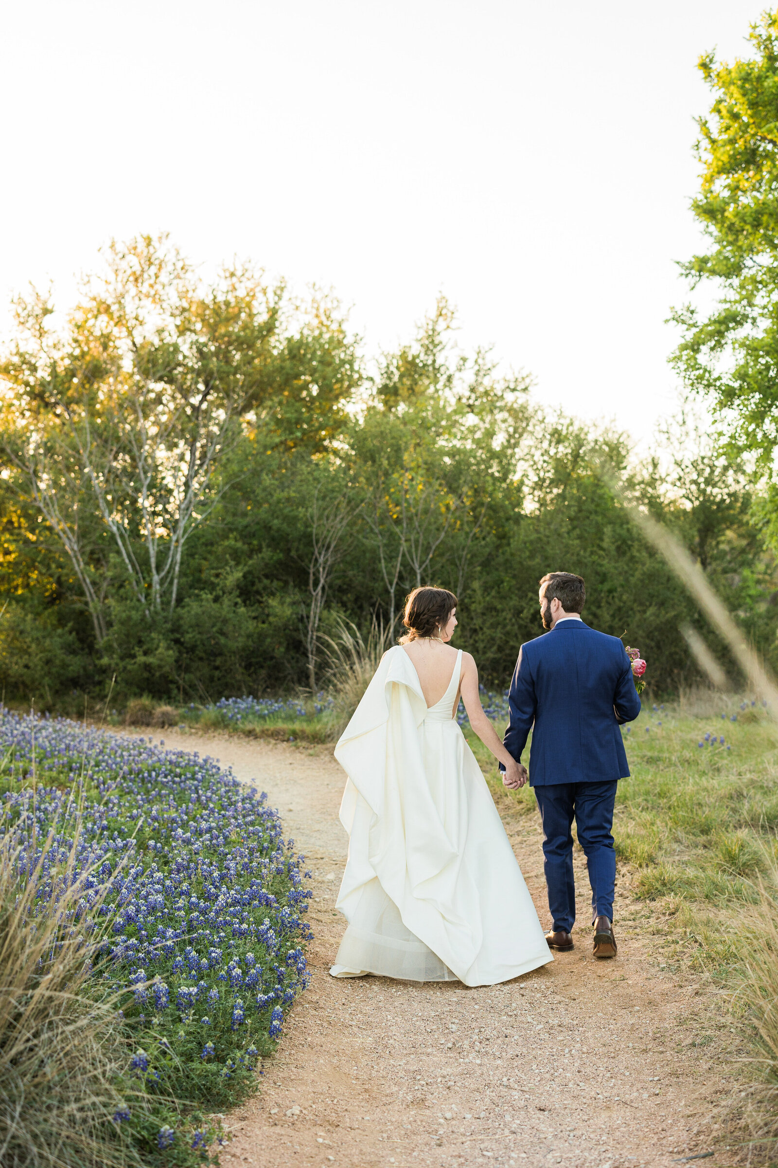 A bride and groom walking down a path away from the camera at the Lady Bird Johnson Wildflower Center in Austin, Texas. The bride is on the left and is wearing a flowing white dress. The groom is on the right and is wearing a blue suit with brown shoes and is holding a bouquet. They both are on a gravel path and are surrounded by wildflowers and trees.